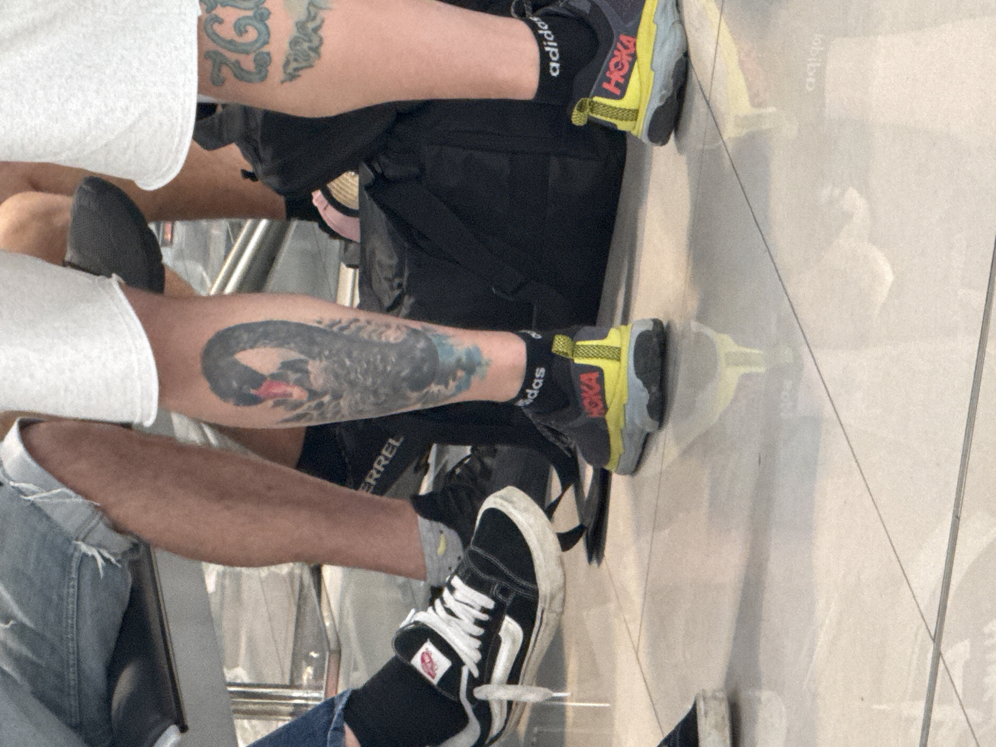 The image captures a low-angle, ground-level view of the lower legs and feet of two individuals, likely seated. The floor appears to be tiled, reflecting light.
The individual on the left has a black Vans sneaker with white laces and a white side stripe. They are wearing a black sock. Their leg is exposed, showing a tattoo of a black swan on the calf. They are wearing denim shorts.
The individual on the right is wearing a dark athletic shoe with a neon yellow accent and a grey sole. They are wearing a black sock with white "adidas" text. Their leg features a tattoo that includes light blue lettering, possibly "ZG".
A black duffel bag is visible behind the individuals' legs. The overall impression is of people waiting or resting, possibly in a public space like an airport or train station. The scene does not provide any direct clues to its specific location within Bratislava, Slovakia.