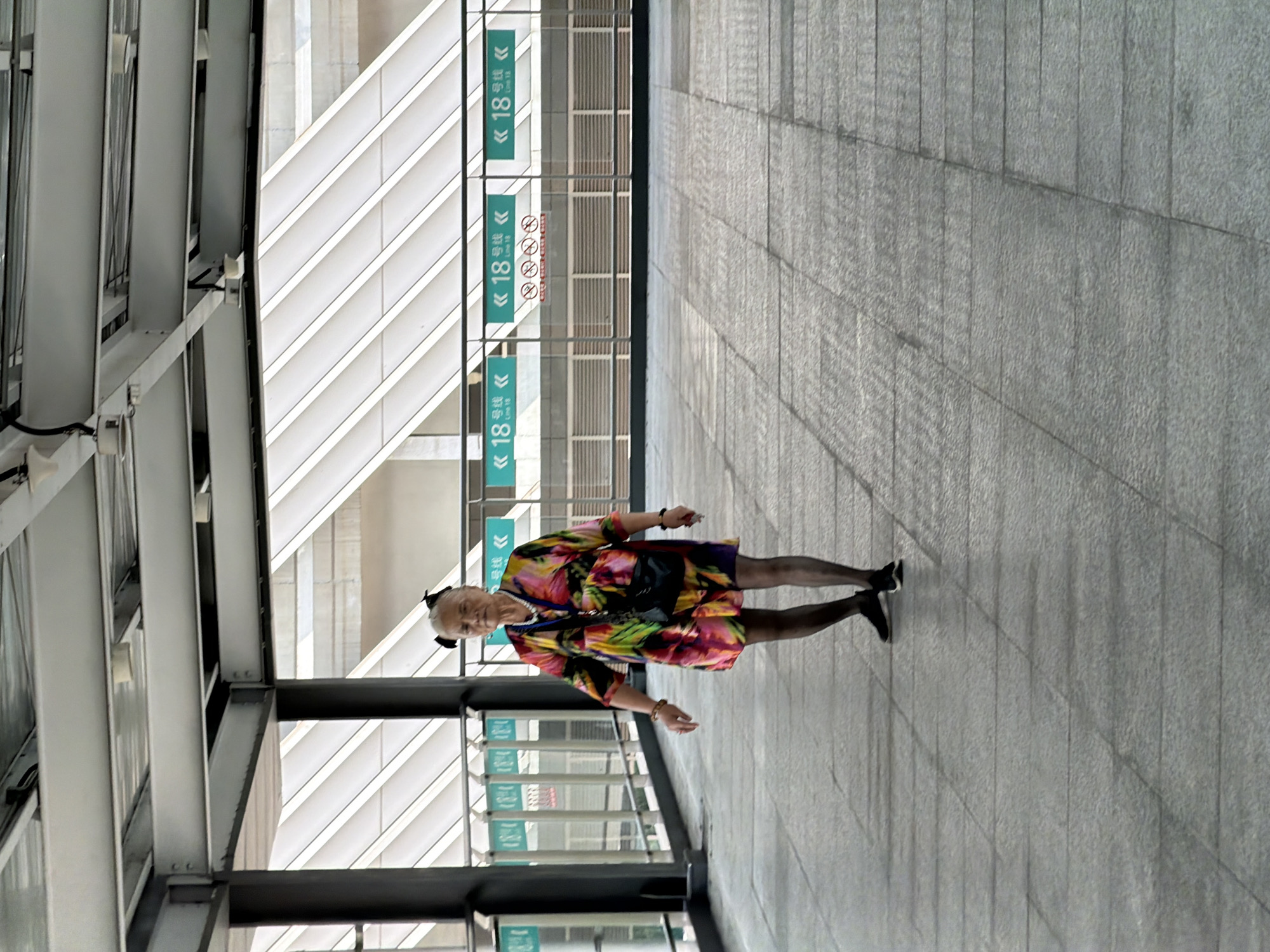 An elderly woman with a black hair bow stands on a tiled pedestrian walkway within a covered outdoor structure. She is wearing a brightly colored, short-sleeved dress with a floral or abstract pattern, sheer black pantyhose, and black athletic shoes. A dark-colored crossbody bag is slung over her right shoulder. She is looking towards the camera. The walkway has a gray tiled floor with a subtle texture. Behind her, a metal railing separates the walkway from a lower level, where signs indicate "Line 18" in green. The structure has a metal ceiling with exposed beams and supports, and translucent panels running diagonally overhead, suggesting a modern urban transportation hub or covered public space in Tiaodeng, China. The lighting is bright and diffused.