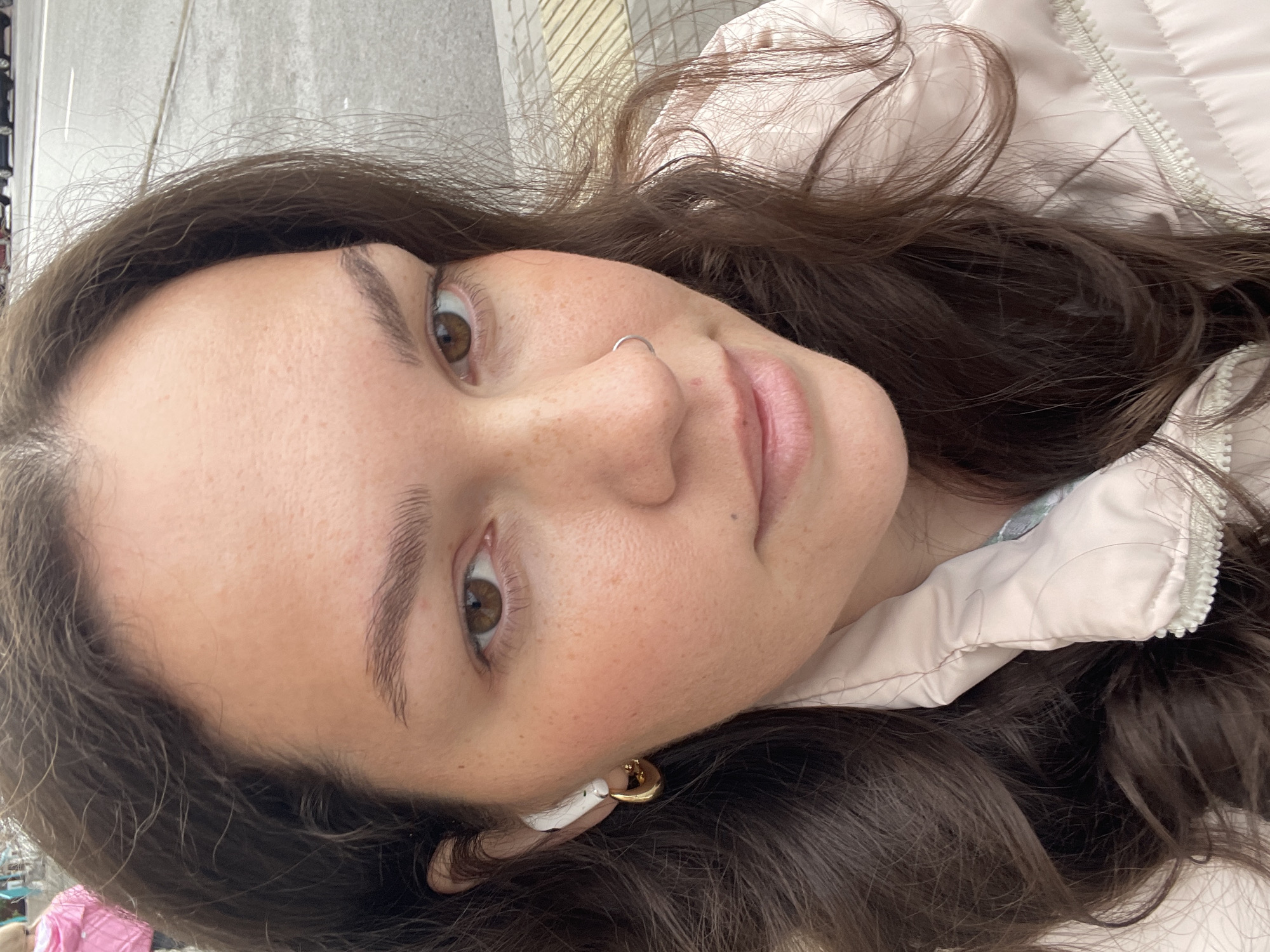 A close-up, slightly tilted selfie of a woman with brown hair and hazel eyes. She has fair skin, freckles, and a silver nose ring in her left nostril. An earbud is visible in her left ear. She is wearing a light pink jacket. The background is indistinct, with a gray concrete-like surface and a hint of yellow. The location in Izhevsk, Russia, cannot be determined from the image alone.