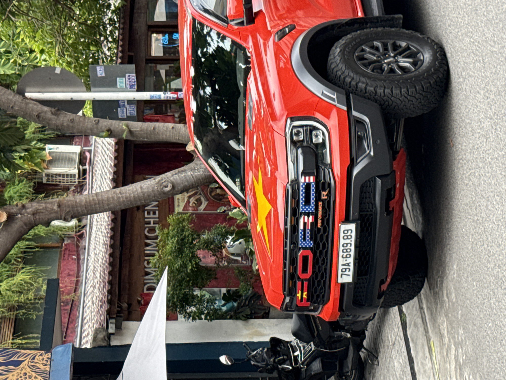 A bright red Ford Raptor truck is parked on a city street in Nha Trang, Vietnam. The truck has a large yellow star emblem on its hood and the word "FORD" emblazoned across its grille, with some letters stylized to resemble the American flag and the Vietnamese flag. The license plate reads "79A-689.89". To the left of the truck, a black motorcycle is parked. Behind the truck, a building with a sign that reads "DIMSUM CHEN" is visible, along with trees, foliage, and other street-level structures. The overall scene depicts an urban environment with a focus on the prominently decorated truck.