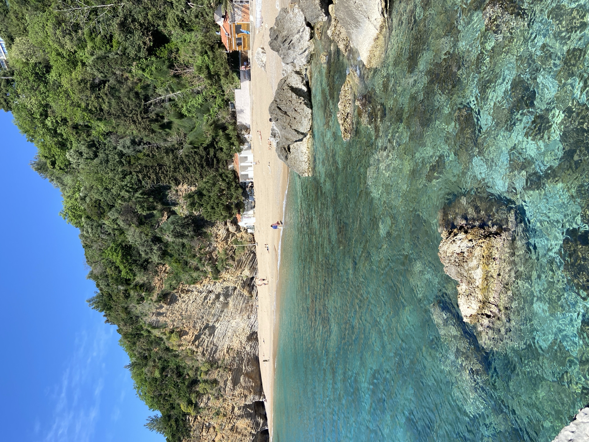 This aerial view captures a picturesque beach cove in Budva, Montenegro, under a clear blue sky. The scene is dominated by the vibrant turquoise and emerald hues of the shallow, crystal-clear sea, revealing the sandy bottom and submerged rocks.
Along the water's edge, a narrow strip of golden sand forms the beach, where several people are seen enjoying the day. Some individuals are visible sunbathing or relaxing on the sand, while others are walking near the shoreline. Small groups are scattered across the beach, suggesting a leisurely atmosphere.
Behind the beach, the terrain rises sharply into steep, rugged cliffs composed of stratified rock formations. These cliffs are densely covered with lush green Mediterranean vegetation and trees, extending to the horizon on the left. Nestled at the base of these cliffs, along the back of the beach, are a few small, modest buildings with red-tiled roofs, possibly beachside cafes or accommodations, adding a touch of human presence to the natural landscape. One small boat is faintly visible near the buildings in one of the crops.
The time of day appears to be bright daylight, possibly late morning or early afternoon, indicated by the strong sunlight illuminating the scene and the minimal cloud cover. The weather is sunny and clear, contributing to the inviting appearance of the water. Large boulders are scattered both on the beach and in the shallow water, adding texture and natural elements to the coastal scene. No discernible text is visible within the image.