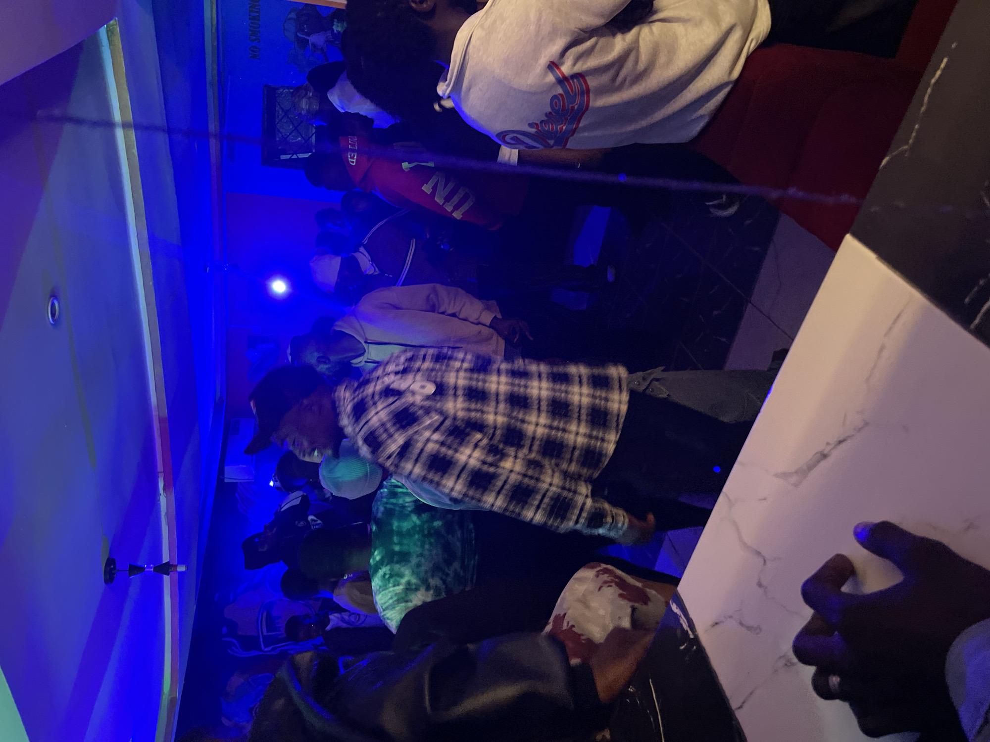 The image depicts a dimly lit indoor venue, possibly a club or bar, in Durumi, Nigeria. The atmosphere is energetic with a group of people gathered, illuminated by vibrant blue and pink lighting. The ceiling features a decorative light fixture. A mural with graffiti-style artwork adorns one wall, with a "NO SMOKING" sign visible.
In the foreground, a person's hand is visible resting on a white marble-patterned surface, likely a bar counter. To the right, a person in a light-colored hoodie is seen from behind. In the center, a person in a plaid shirt is facing away from the camera, seemingly engaged in the activity. Other individuals are scattered throughout the scene, some wearing hats or hoodies, suggesting a casual social gathering. The overall impression is one of a lively night out.