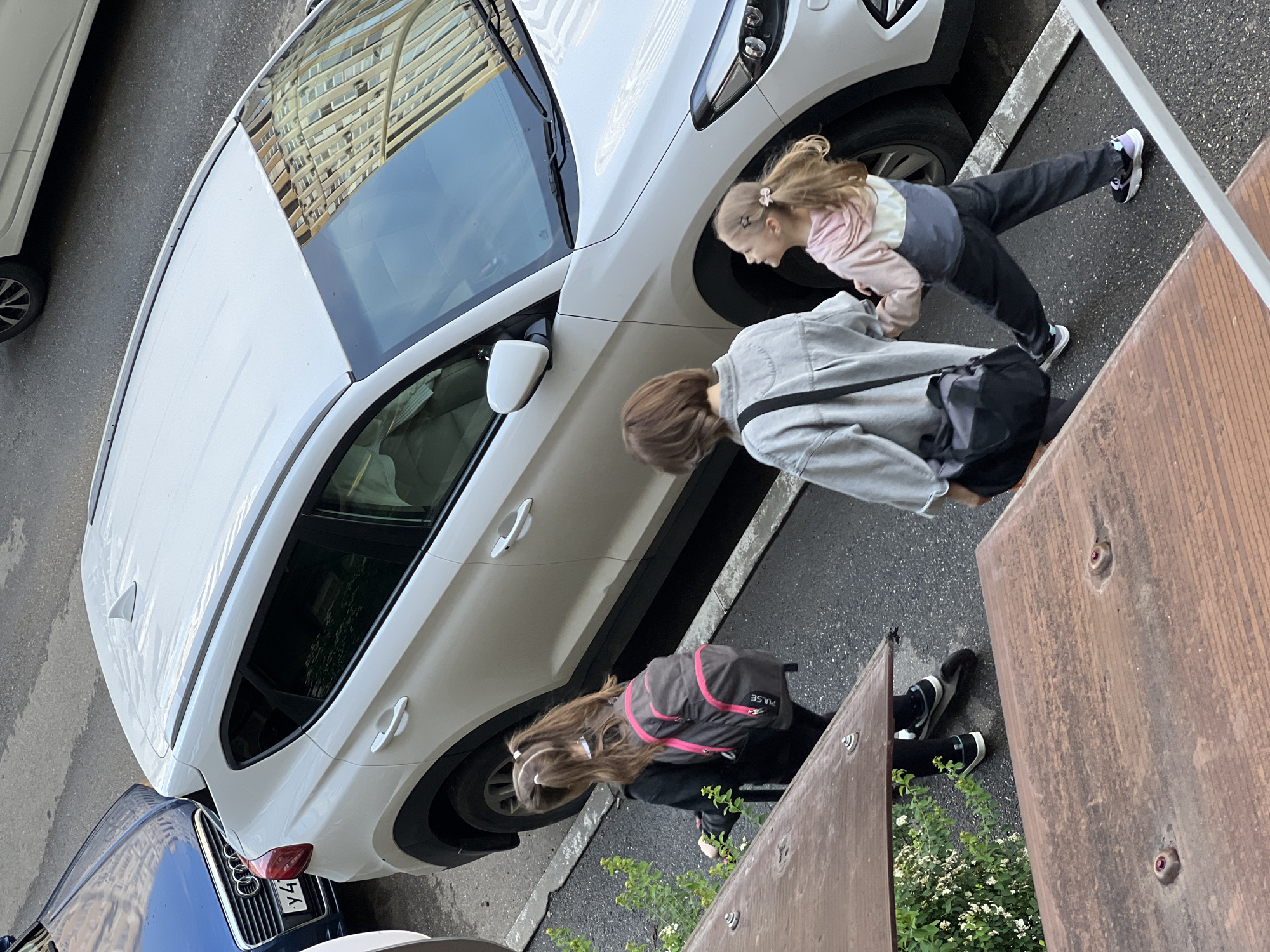 The image depicts an outdoor scene in a residential area. A white Mercedes SUV is parked on the street next to a blue Audi, both oriented towards the lower left of the frame. Three girls are present; one wears a pink and grey top, another a grey jacket with a black bag, and the third a black top with a pink and grey backpack. They are standing next to a wooden fence or barrier, engaging in some sort of interaction or activity. Some green plants and flowers are visible growing near the fence. The scene is situated in Krasnodar, Russia.