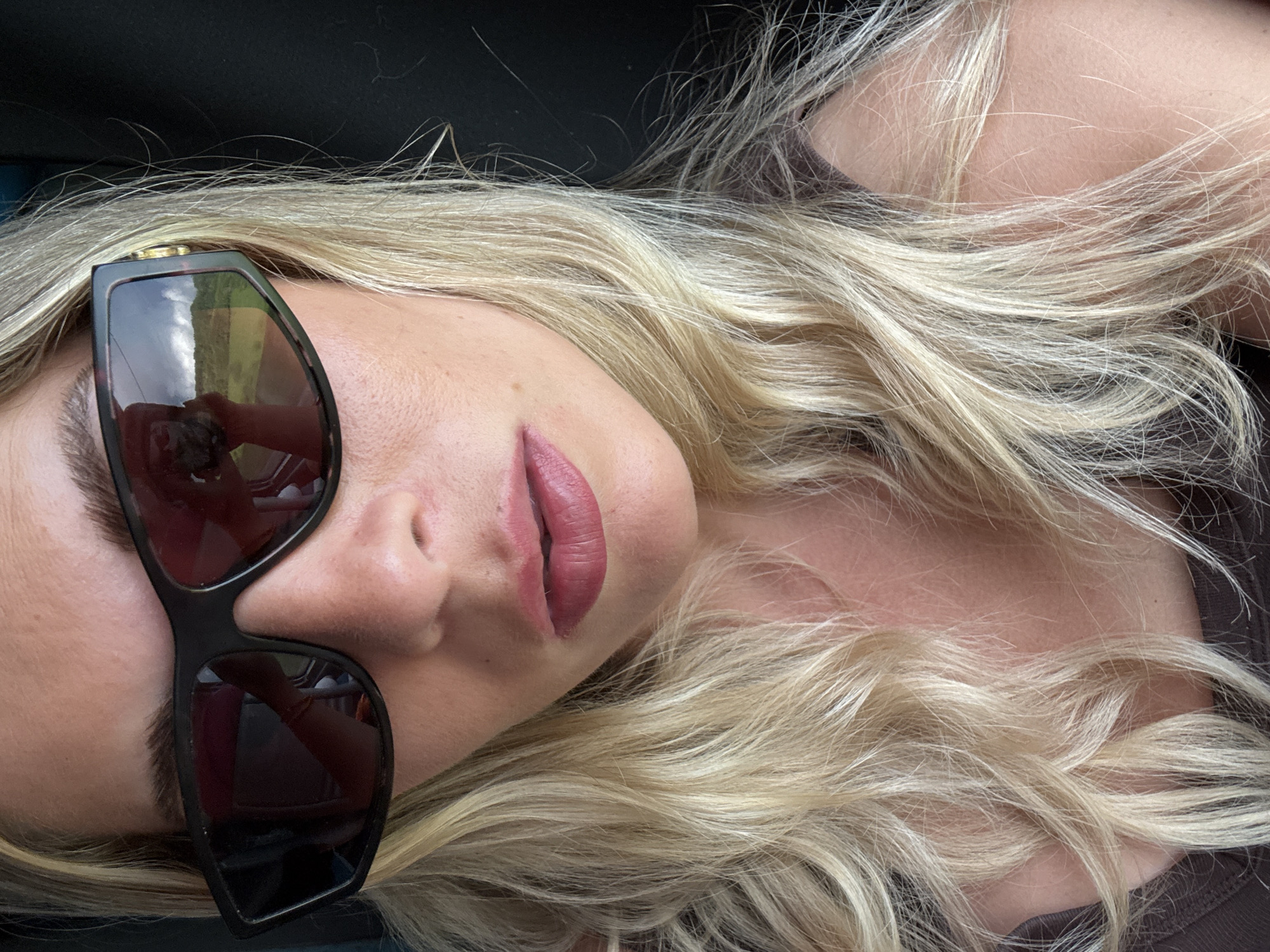 A light-skinned woman with blonde hair is reclining, possibly outdoors in or near Lournand, France. She is wearing sunglasses reflecting greenery and possibly a building, and is wearing a dark top. The angle is looking down at her from above and slightly to the side. The image is well lit. Her lips are a deep pink/mauve color.