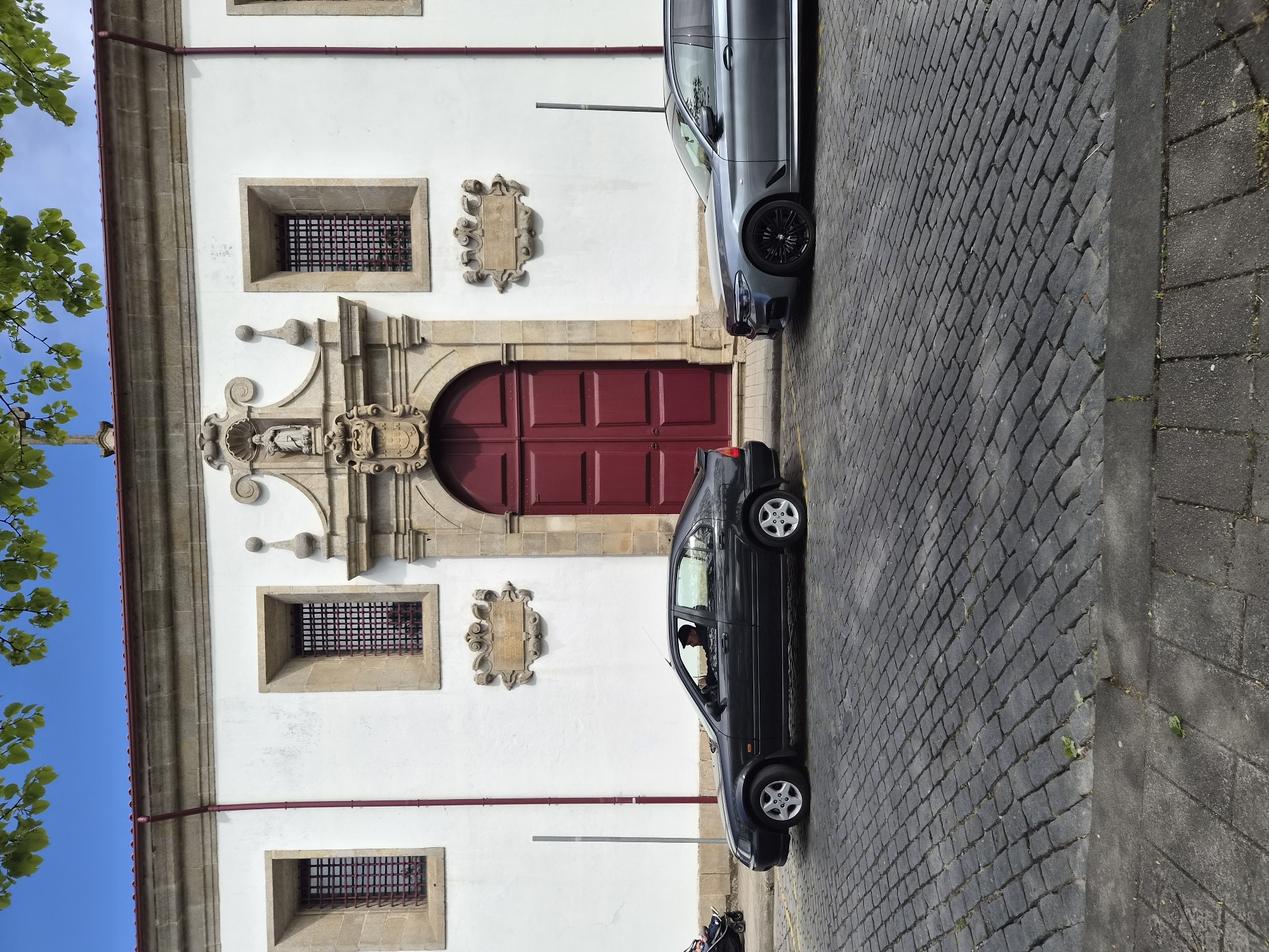 The image shows a white building in Barcelos, Portugal, with a red arched door, barred windows, and stone carvings. Two cars are parked on the cobblestone street in front of the building: a silver car and a black car with a person visible in the driver's seat. Part of a tree is visible in the upper left of the frame.