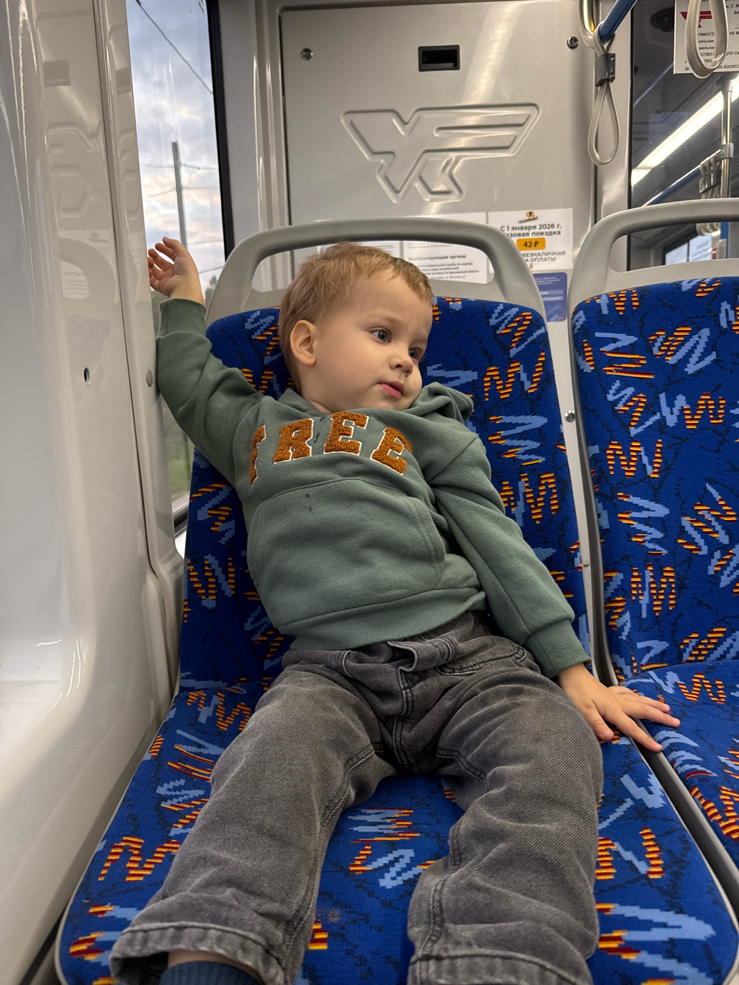A young child with blonde hair sits on a blue patterned seat on a public transport vehicle. The child is wearing a light green hooded sweatshirt with "FREE" written on it in orange lettering and grey pants. The child's right arm is raised and resting against a white surface. The background shows the interior of the vehicle, including another blue patterned seat and a window. There are some signs on the wall of the vehicle, one with the Cyrillic text "42 Р". The image appears to have been taken in Ignatovo, Russia, based on the provided context.