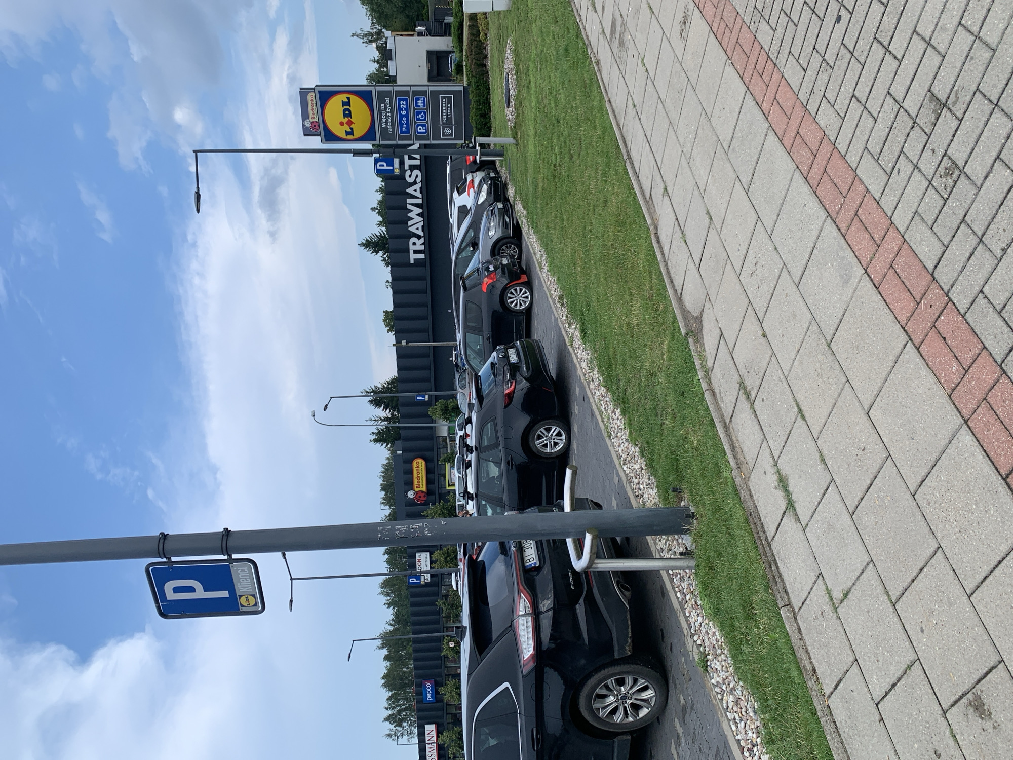 The image shows a parking lot outside a shopping center in Białystok, Poland. The shopping center has several stores, including Lidl and Biedronka. The parking lot is full of cars, and there are several signs indicating parking for customers. The weather is partly cloudy. The image is taken from a slightly elevated perspective, showing the parking lot, the shopping center, and the surrounding area.