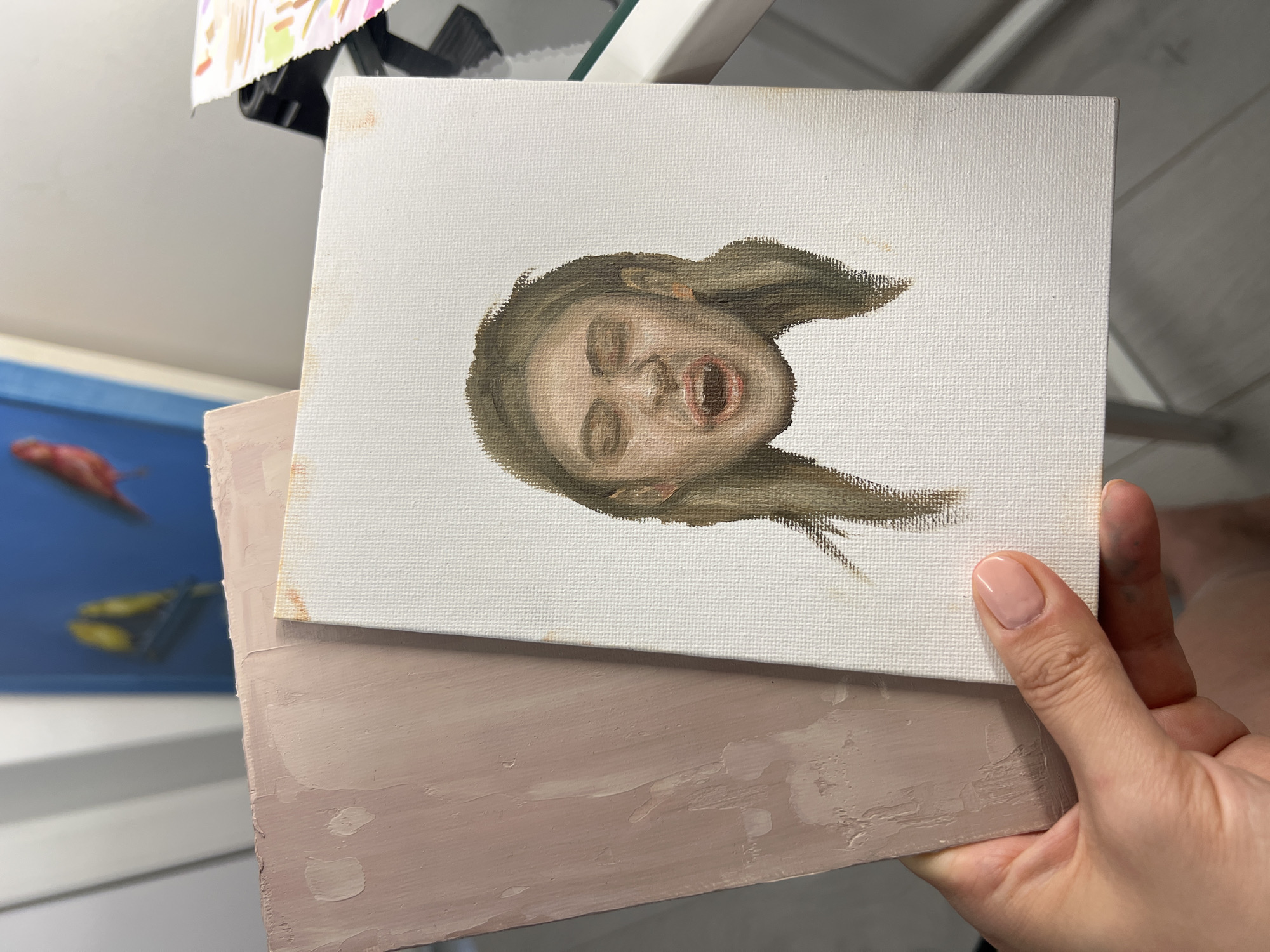 In the foreground, a hand with pale pink nail polish holds two art canvases. The canvas on the right, smaller and positioned slightly forward, features a nearly completed oil painting of a woman's face with her mouth open in a scream or yell. Her eyes are closed, and her hair is a medium brown, falling loosely around her face. The canvas has a few small orange smudges on its white surface. To the left of this canvas is a larger, thicker canvas or board, coated in a thick, textured pinkish-beige paint with visible brushstrokes. The background is out of focus, showing hints of a blue wall with what appear to be painted birds, a white surface, and some colorful papers. The setting appears to be an indoor studio or workspace.