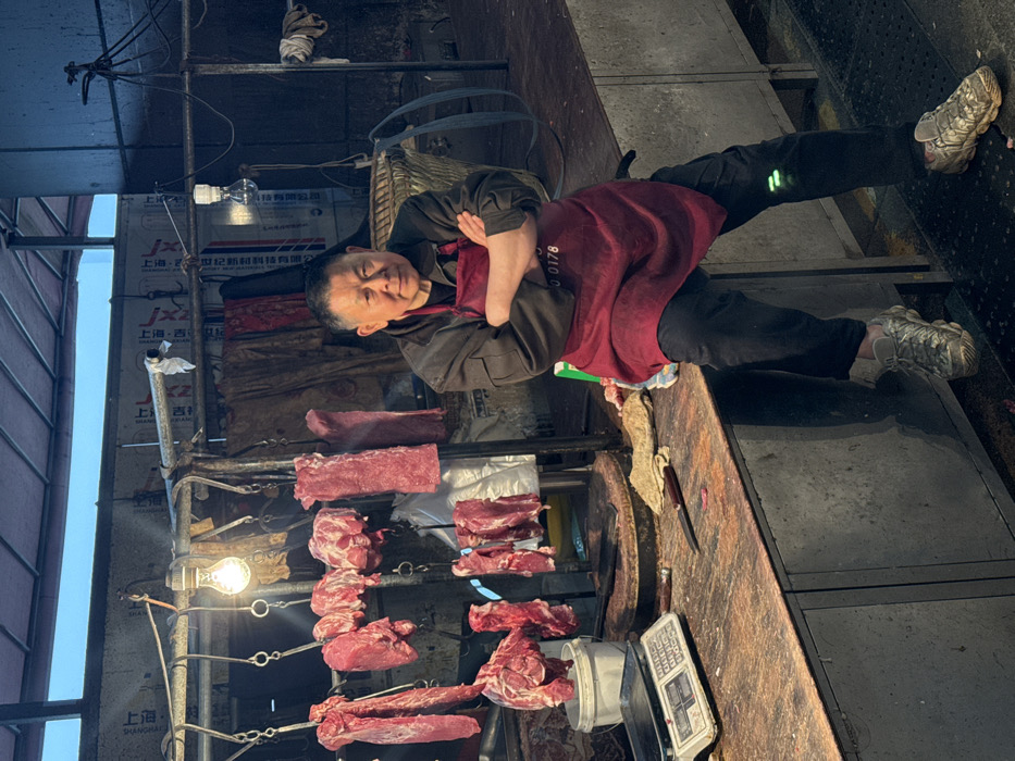 An East Asian male, appearing middle-aged to elderly, is depicted sitting on a dark metal counter within a market stall in Jiangjin, China. He is wearing a dark jacket, a dark red apron with the visible numbers "0178," black trousers, and grey/camouflage patterned athletic shoes. His arms are crossed over his chest, and he gazes directly forward.
To his left, on the counter, a white digital scale with a metal platform is present. Next to it, a well-worn circular wooden cutting block holds a dark-handled cleaver, with another knife resting directly on the counter. A crumpled light-colored cloth and small pieces of raw meat are also on the counter.
Above and behind the man, numerous raw red meat cuts of varying sizes hang from metal hooks suspended from a scaffold-like structure. A single bare incandescent light bulb, actively illuminated, hangs from this structure. The background consists of dark corrugated metal panels, some displaying Chinese characters such as "上海" and other smaller text. A large, dark woven basket is visible behind the man to his right. A white plastic bucket is situated beneath some hanging meat on the left. The upper background shows an expanse of light blue sky. The floor beneath the counter is dark and textured.