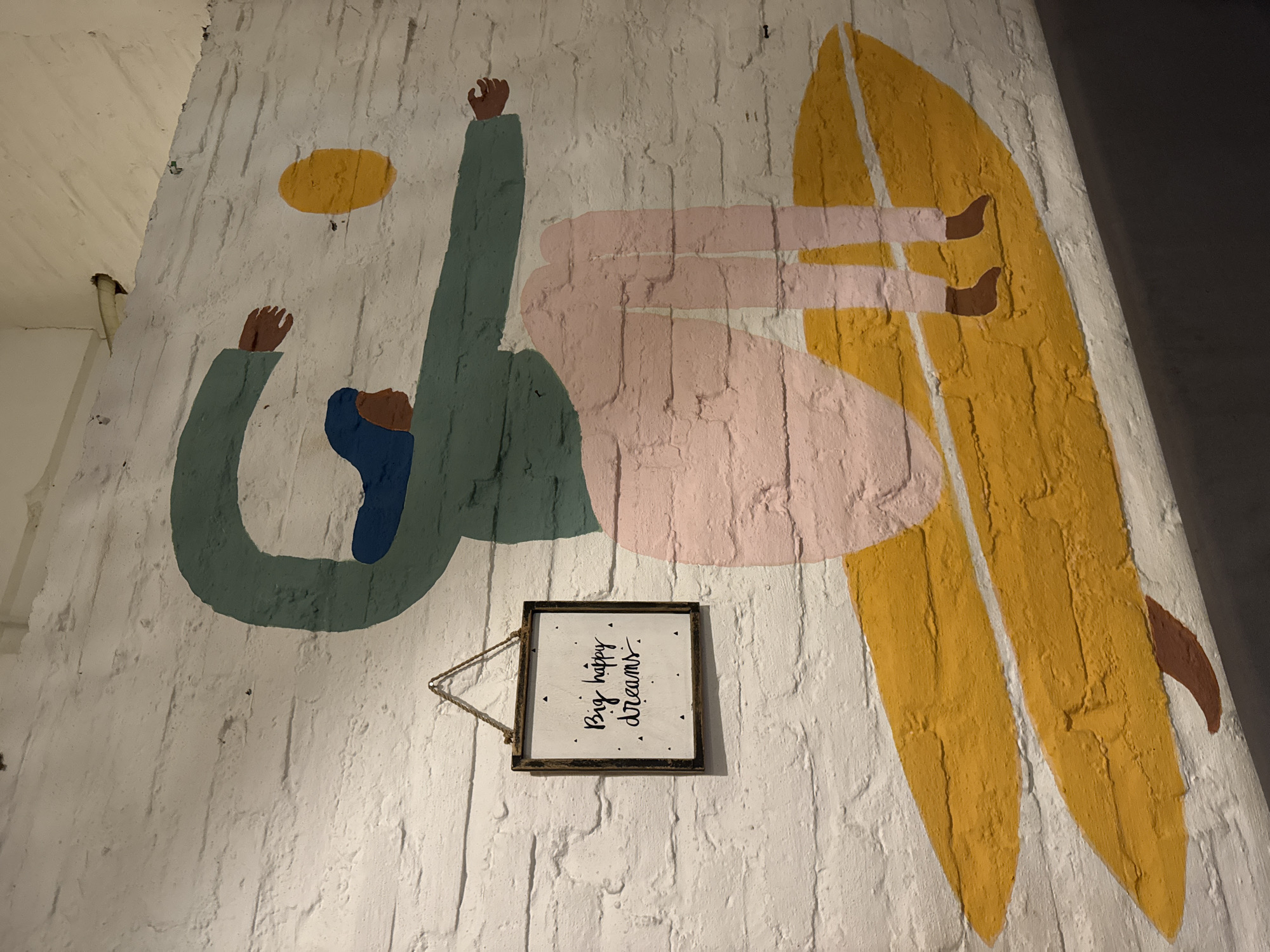 This media file captures a vibrant mural painted on a textured white brick wall, likely within an indoor or covered space in Kuta, Indonesia. The mural features two stylized human figures rendered in a minimalist, block-color art style. One figure, positioned upright on the left, wears a green top with blue and brown accents and reaches upwards towards an abstract yellow circular shape. The second figure, depicted upside down, wears a light pink outfit and appears to be interacting with a large, yellow surfboard that dominates the right side of the mural. This arrangement suggests a playful, dream-like scene, possibly related to surfing or floating.
Below the mural's figures, a small, dark-framed sign hangs against the white brick wall. The sign displays the handwritten text: "Big happy dreams." The overall lighting is somewhat dim, suggesting artificial light or an evening setting, which emphasizes the bright colors of the mural. The textured brick surface is visible throughout the image, giving depth to the painting. The combination of surfing imagery and the "Big happy dreams" text evokes themes of aspiration, freedom, and the popular beach culture associated with Kuta, Indonesia.