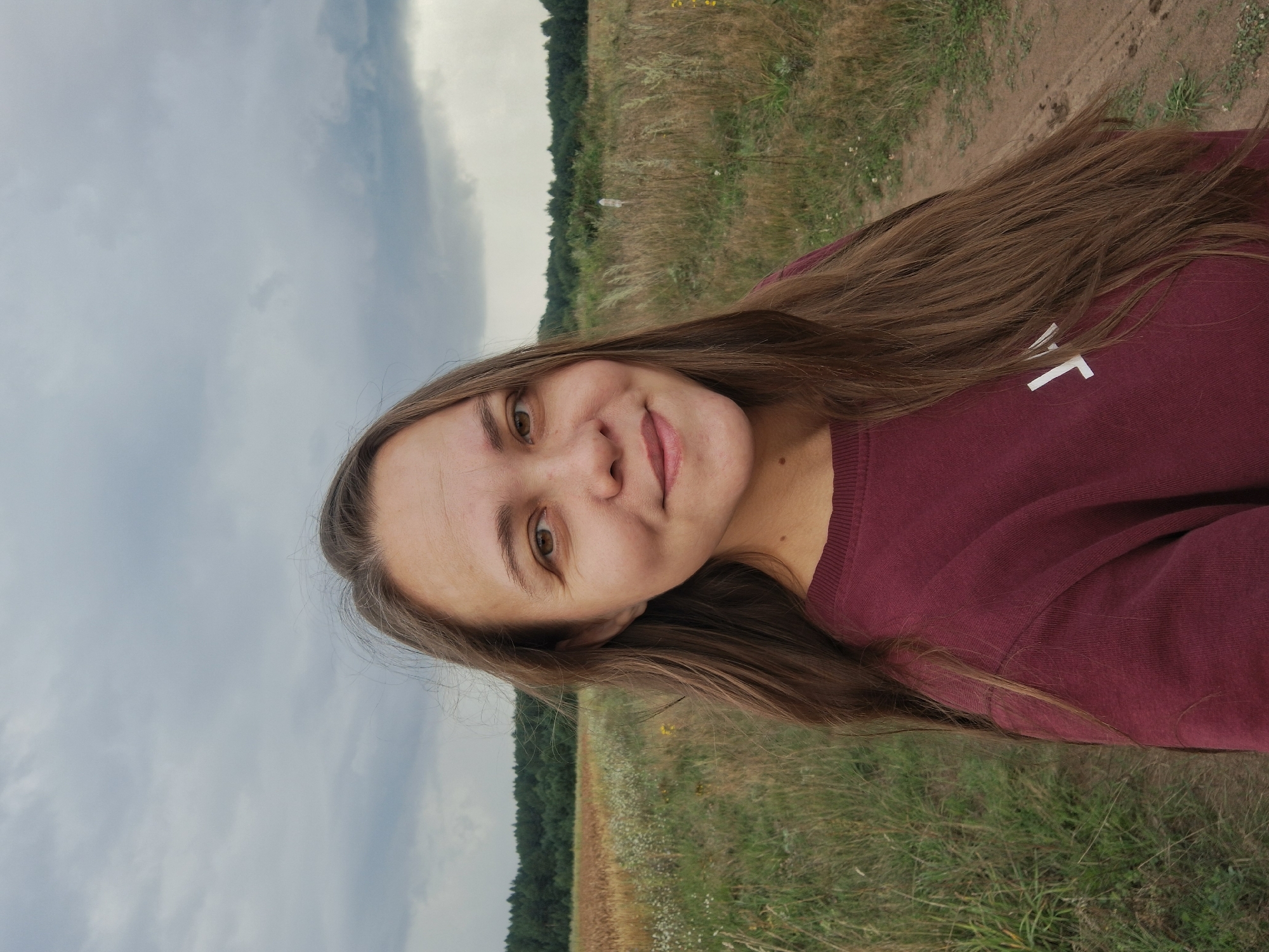 A medium shot captures a woman with long, brown hair pulled to the side and a burgundy sweatshirt, smiling faintly as she looks directly at the camera. She is positioned in the foreground, with a field of tall, dry grass and scattered white wildflowers stretching out behind her. A dirt path is visible to her right, leading into the distance. A dark green forest forms the backdrop to the field. The sky above is overcast with dramatic, gray clouds. The image appears to have been taken outdoors in a rural setting, possibly the outskirts of Plishkino, Russia.
