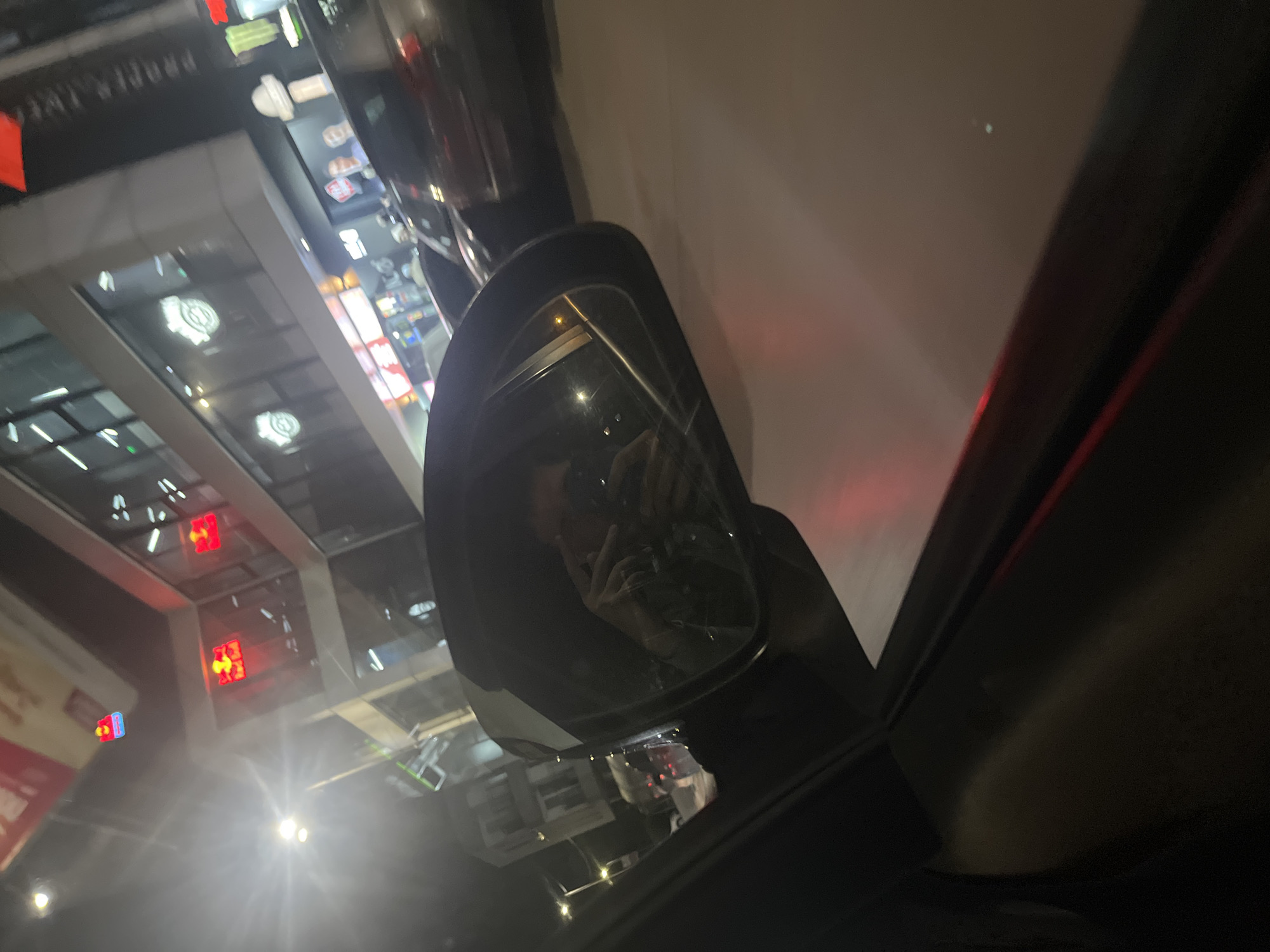The image depicts a car's side mirror, reflecting a nighttime street scene. The reflection includes buildings with illuminated signs, likely storefronts, and streetlights. The perspective suggests the photograph was taken from inside the car. Given the reference to Sofia, Bulgaria, it can be inferred the location is within the city, but without additional landmarks, a precise location is not identifiable. The mirror also reflects a person taking the photograph.