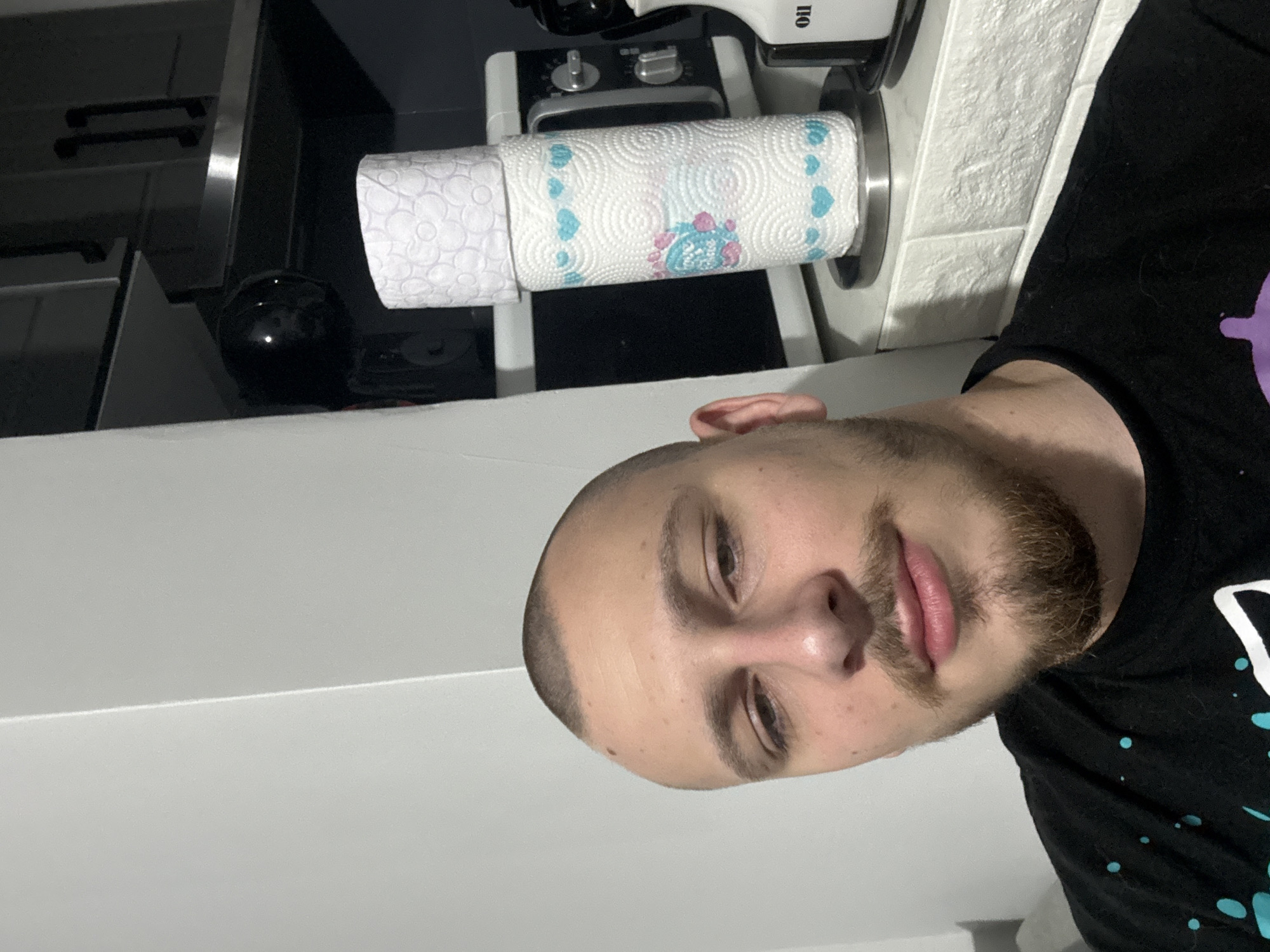 A person is standing in what appears to be a kitchen or bathroom. The person is facing the camera and is close enough that their face is the main subject of the image. The person has short hair and a beard. They are wearing a black t-shirt.
Behind the person, there's a roll of paper towels on a dispenser. There are kitchen cabinets and possibly other kitchen appliances in the background, but they are somewhat out of focus. There is also a black object, possibly a kitchen tool, on the countertop. The background suggests an indoor domestic setting.
The image provides no information about the location being Stara Zagora, Bulgaria.