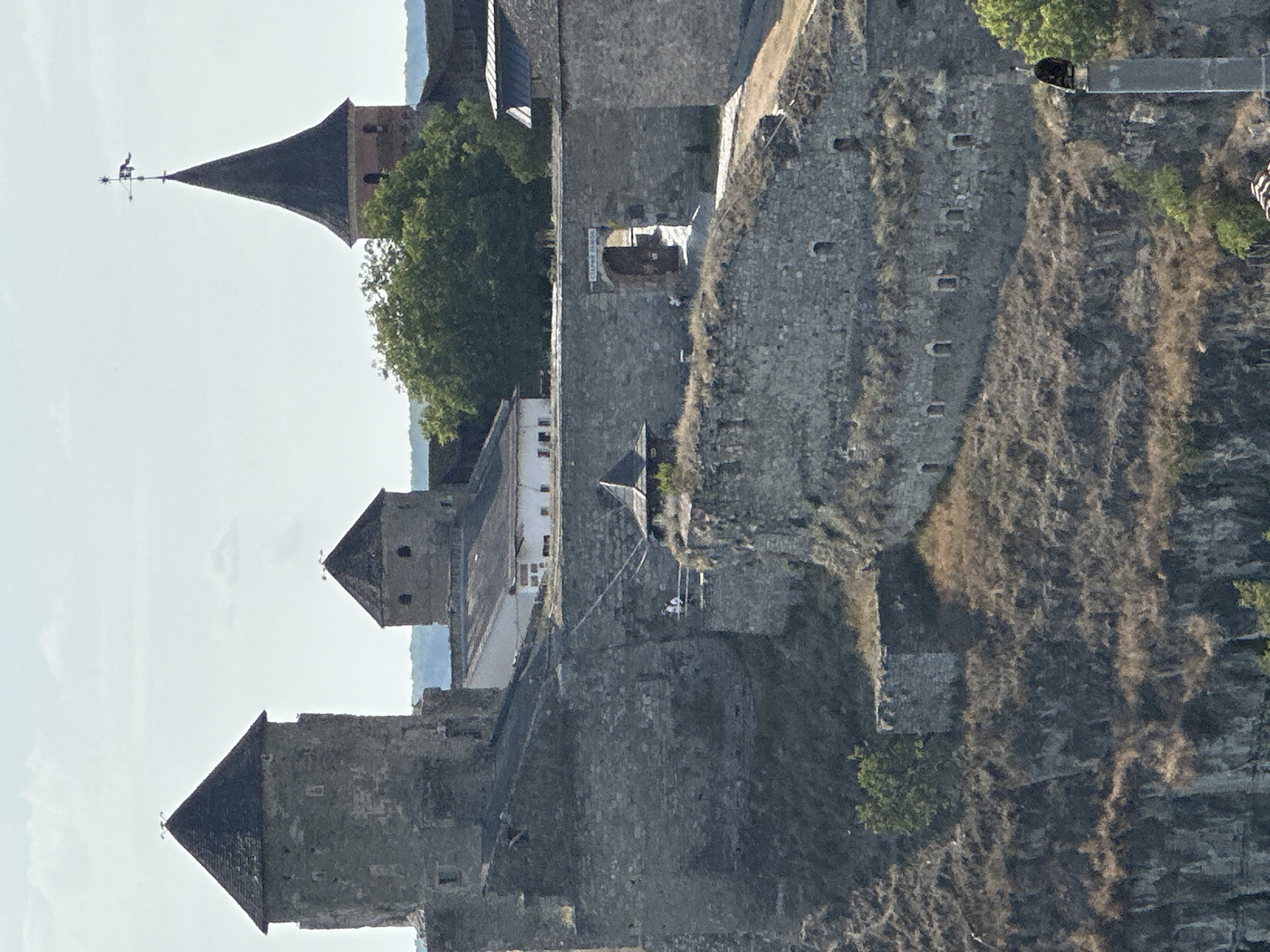 The image captures a view of the Kamianets-Podilskyi Castle in Kamianets-Podilskyi, Ukraine. The imposing stone fortress is situated on a rocky promontory, with steep, overgrown slopes leading up to its walls. Multiple towers, some conical-roofed and others with flat tops, are visible, constructed from large, rough-hewn stones. A prominent tower on the left has a dark, pointed roof, while another in the center is shorter and also features a conical roof. In the background, a tall, slender tower with a dark conical roof is topped with a weather vane.
The castle walls are substantial and show signs of age, with visible stone construction and small arched openings. A wide stone gateway with a wooden door is situated in the middle ground. There are some figures on a walkway near the wall, appearing as small white shapes. A wooden, pitched-roof structure with a dark roof is perched on a rocky outcrop. Vegetation, including dry grass and a few trees, grows on the slopes and within the castle grounds. The sky above is a pale blue with scattered white clouds. In the far right foreground, a tall, grey cylindrical structure, possibly a chimney or part of the fortifications, has a black object perched on its top. The overall scene conveys a sense of historical grandeur and fortification.