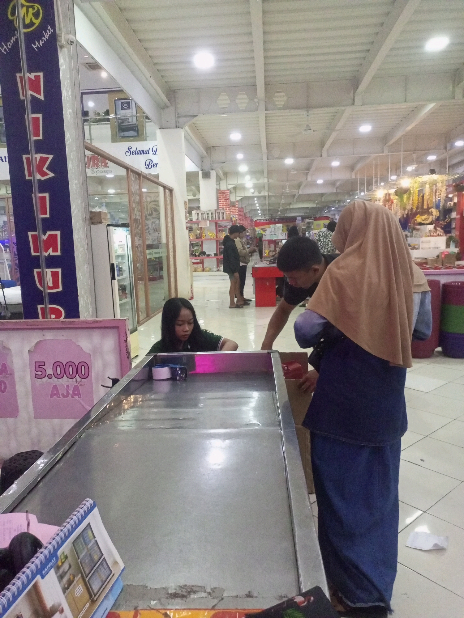 In a commercial indoor setting in Ngasem, Indonesia, two individuals, a woman and a man, are engaged in an activity at a long, metallic table. The woman, with dark hair, is seated and looking downwards, her hands near the table surface. The man stands behind her, leaning over the table, with his hands also positioned on or near it. A third person, wearing a light brown hijab and a dark blue skirt, is standing to the right of the man, partially obscuring him and also looking towards the table. The table appears to be a game or demonstration area, with various items on its surface and a pink sign with text, including "5.000 AJA," visible in the foreground. The environment suggests a market or retail space, with ceiling lights, white walls, and displays of goods visible in the background. A blue banner with white lettering is prominent on the left side of the image.