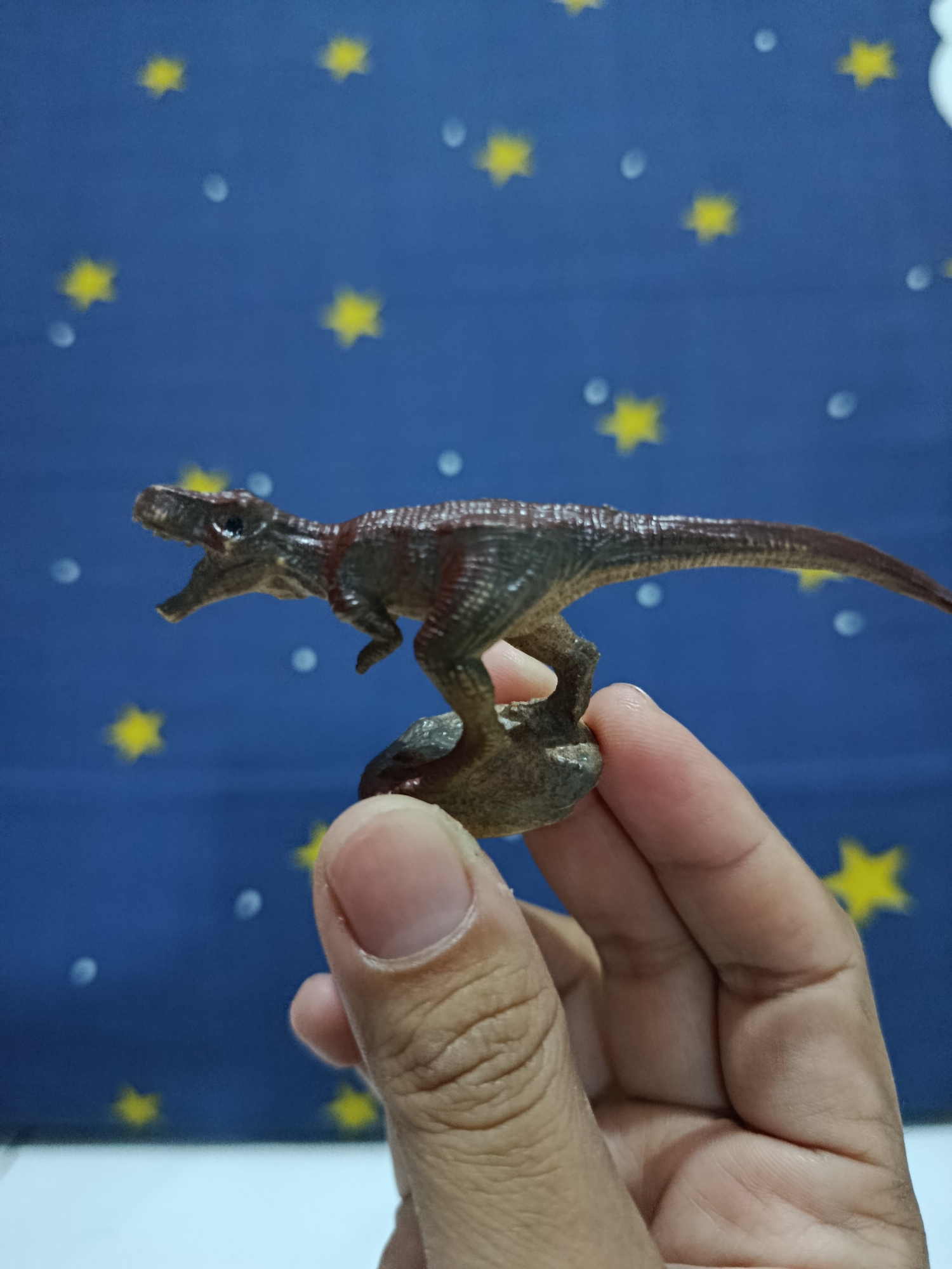 A person's hand is holding a small, brown and green toy dinosaur on a brown base. The dinosaur is posed in a quadrupedal stance with its mouth open and its head turned to the left. The background is a dark blue fabric with yellow stars and white circles. The scene is lit from the front, casting a shadow on the hand. The image was taken in Wanasari, Indonesia.
