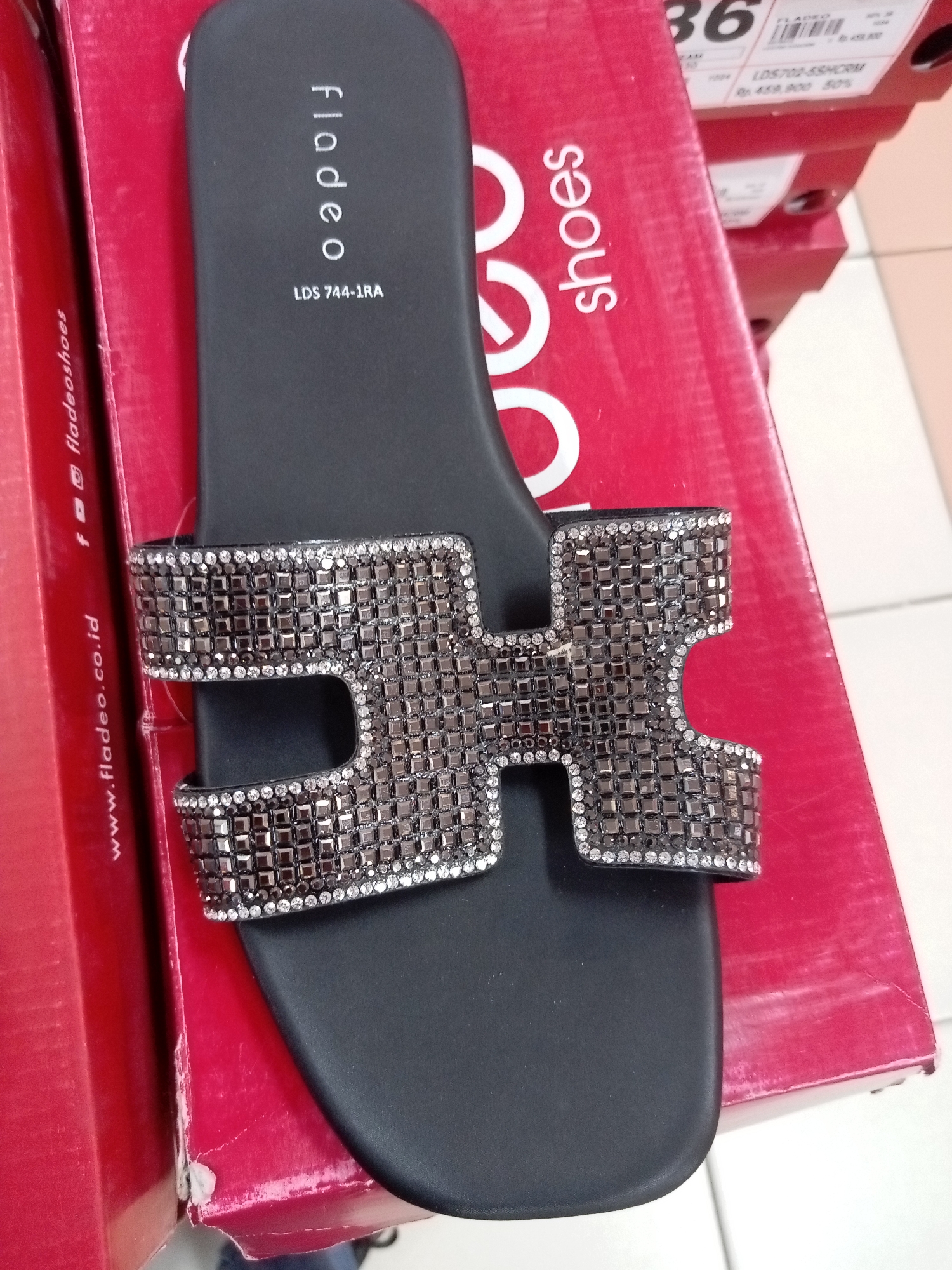 An overhead shot shows a black slide sandal with an H-shaped strap adorned with dark and clear rhinestones. The sandal is placed on top of a red shoebox with the text "fladeo shoes" printed in white on its side, along with a website address "www.fladeo.co.id". The shoebox is part of a stack of similar red shoeboxes. In the background, other shoeboxes are visible, and a price tag indicating "Rp 459,900 50%" and size "36" is attached to one of them. The image appears to be taken in a retail setting, possibly a shoe store in Batam, Indonesia.