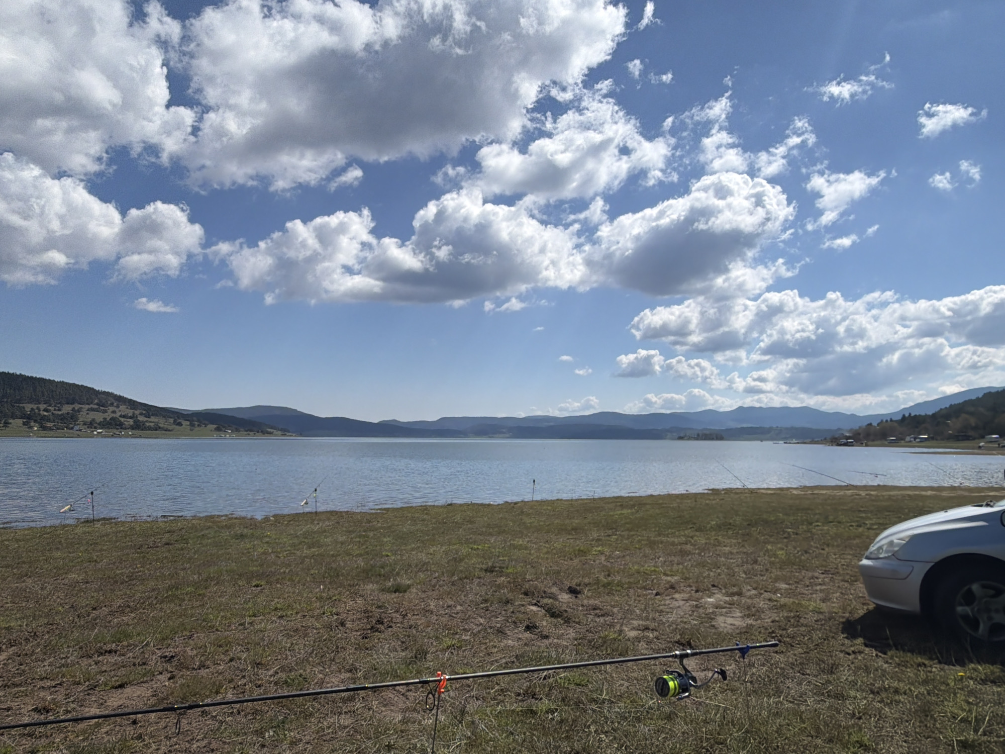 The image depicts a scenic view of a lake near Rakitovo, Bulgaria, under a bright blue sky with fluffy white clouds. In the foreground, there's a grassy area with a fishing rod and reel laid out. Other fishing rods are also placed along the shoreline, indicating fishing activity. A silver car is parked on the right side of the frame. Across the lake, there are rolling hills and mountains in the distance.