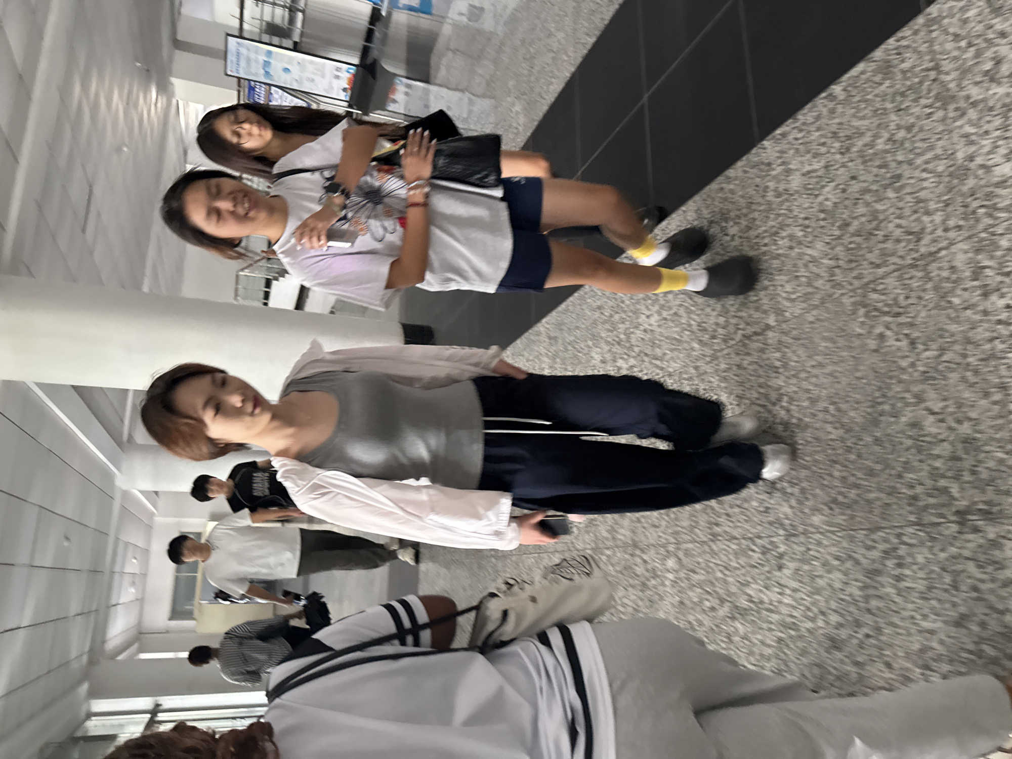 A group of young people, likely students, are walking down a well-lit indoor hallway. The hallway has a speckled gray floor and white pillars. On the right side of the hallway, there is a display board with some text and images. The individuals are dressed casually in t-shirts, shorts, and pants. One person in the foreground is wearing a gray tank top and dark pants, with a light-colored jacket draped over their shoulders. Another person is wearing a white t-shirt with a graphic design and dark shorts, paired with yellow socks and black shoes. The image appears to be taken in a school or university setting, possibly within Guta, China, given the presence of educational posters.