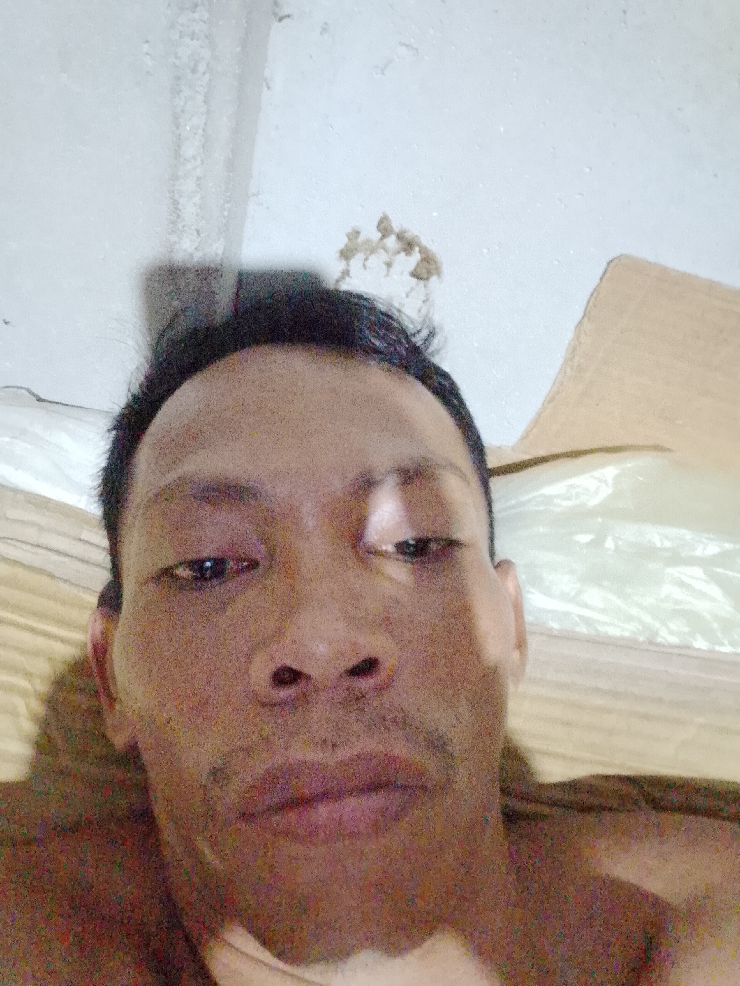 The image shows a close-up selfie of an adult male with short black hair. He is looking down with a neutral expression. The background appears to be an indoor setting, with a white wall and cardboard boxes partially visible. The location is Bukit Ketok, Indonesia.