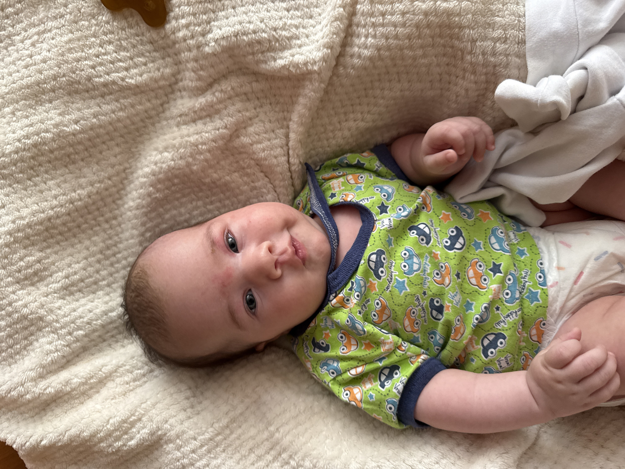 A baby is lying on a cream-colored, fuzzy blanket. The baby is wearing a green short-sleeved onesie with a pattern of cars and stars. The baby has a diaper on and is partially covered by a white cloth. The baby's face is turned slightly to the right, and they appear to be looking at the camera. There are some small red bumps on the baby's forehead. The background is out of focus, but it appears to be the same fuzzy blanket. There is a small, brown object, possibly a toy, to the right of the baby's head. The image is taken from a top-down perspective. The scene is indoors.