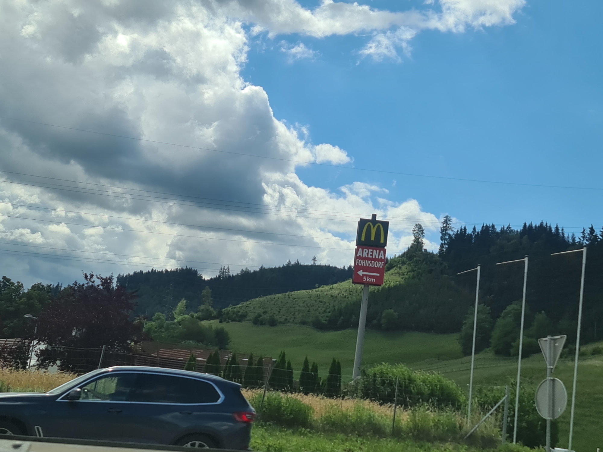 The image shows a road scene with a dark colored SUV in the foreground. In the background, a McDonald's sign is visible, indicating "ARENA FOHNSDORF" and a direction with "5 km". The sign is placed on a pole in front of a hillside covered with green grass and trees. The sky is blue with scattered white clouds. The overall environment appears to be rural or suburban, with indications of a roadside location. Based on the provided information, this scene is located in or near Judenburg, Austria.