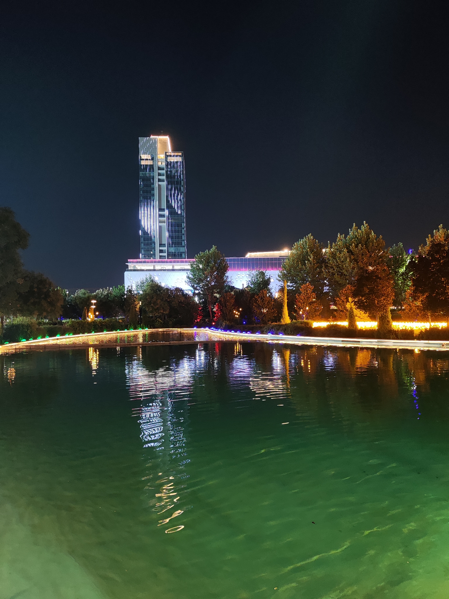 This is a nighttime photograph of the International Business Center in Tashkent, Uzbekistan, specifically the Tashkent City Park area. The central subject is a tall, modern skyscraper illuminated with bright white and yellow lights, featuring vertical LED strips. In the foreground, a calm body of water, appearing green due to lighting, reflects the lights from the buildings and decorative lighting along a pathway or bridge. Trees with illuminated foliage and decorative lights line the far bank of the water. The sky is dark, indicating nighttime. There are no people visible in the frame. The overall impression is one of an urban landscape at night, showcasing modern architecture and park illumination.