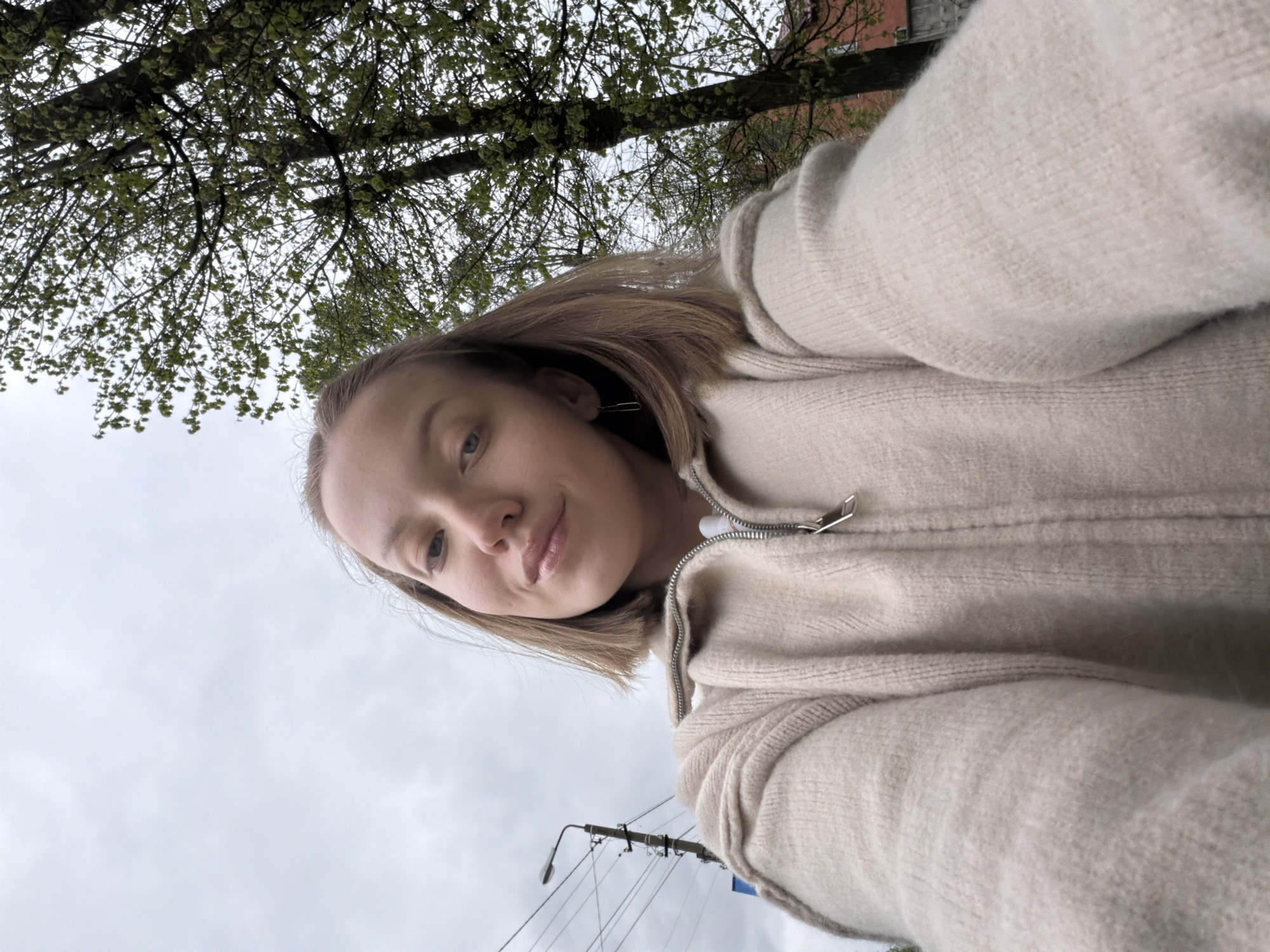A young woman is taking a selfie outdoors. She is wearing a light-colored, zipped sweater. Above her head are tree branches with green leaves and a cloudy sky. In the background, there's a street lamp. Based on the provided information, this photo could be taken in Reshetnikovo, Russia. The focus is on the woman, with the background suggesting an urban or suburban setting.