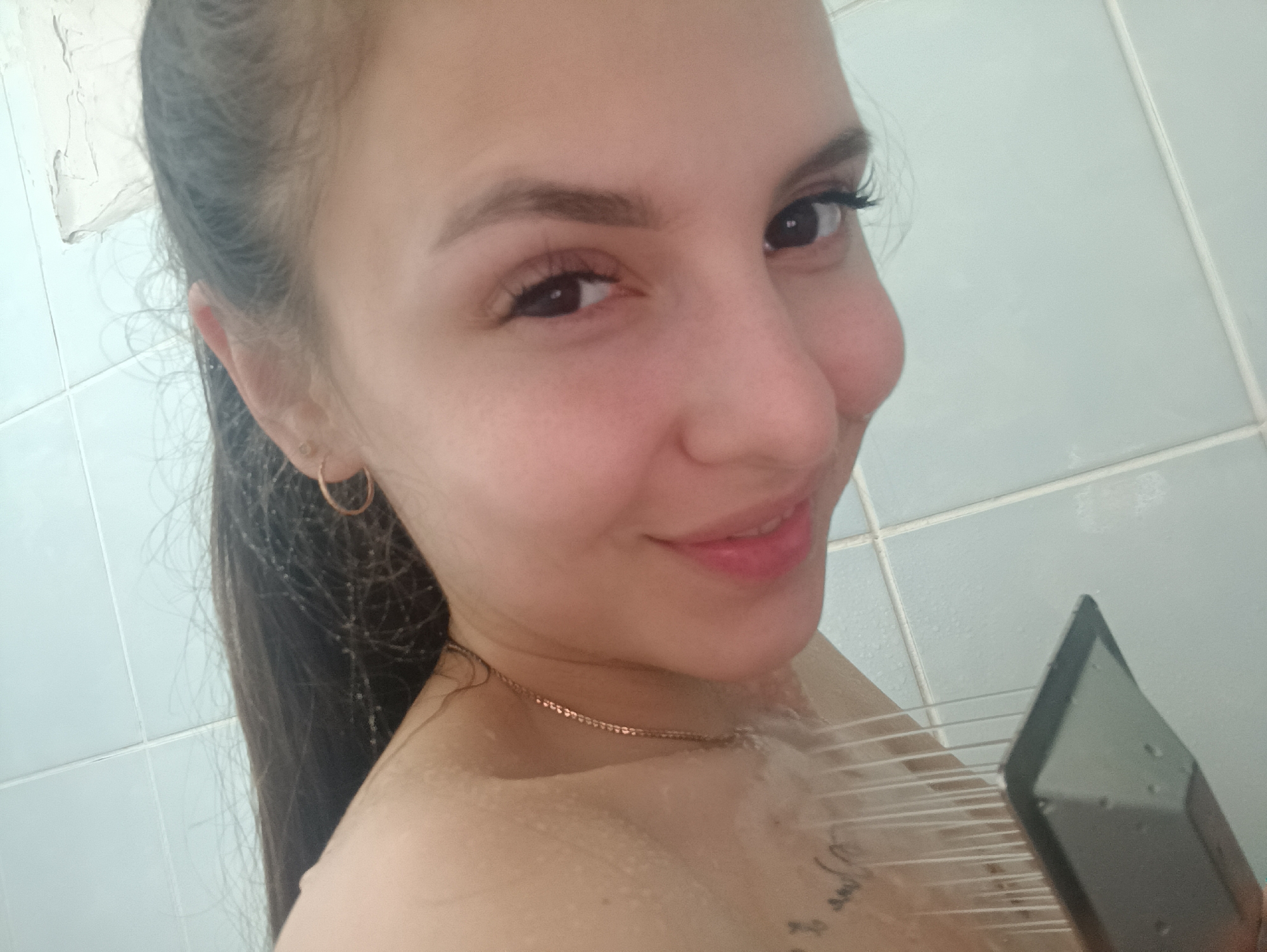 A close-up shot features a woman with long, dark hair pulled back in a ponytail, wearing a gold hoop earring and a gold chain necklace. She is in a shower, evident by the water streaming over her bare skin and the presence of a squeegee in her hand. The shower walls are tiled with light-colored, rectangular tiles. The location appears to be Unknown, Unknown.