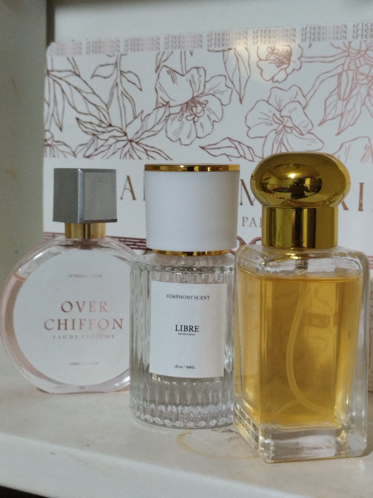 The image shows three bottles of perfume sitting on a white surface. The leftmost bottle is round with a silver cap and a label that reads "AFTER SKIN OVER CHIFFON EAU DE PARFUME." The center bottle is cylindrical with a white cap and gold trim and a label that reads "SYMPHONY SCENT LIBRE 1 fl oz / 30ML". The rightmost bottle is rectangular with a gold, rounded cap. Behind the perfume bottles is a white background with floral designs. The text "AFTERMY SKIN" is visible on the background. The location and city of Lhokseumawe, Indonesia, are not discernible from the image.