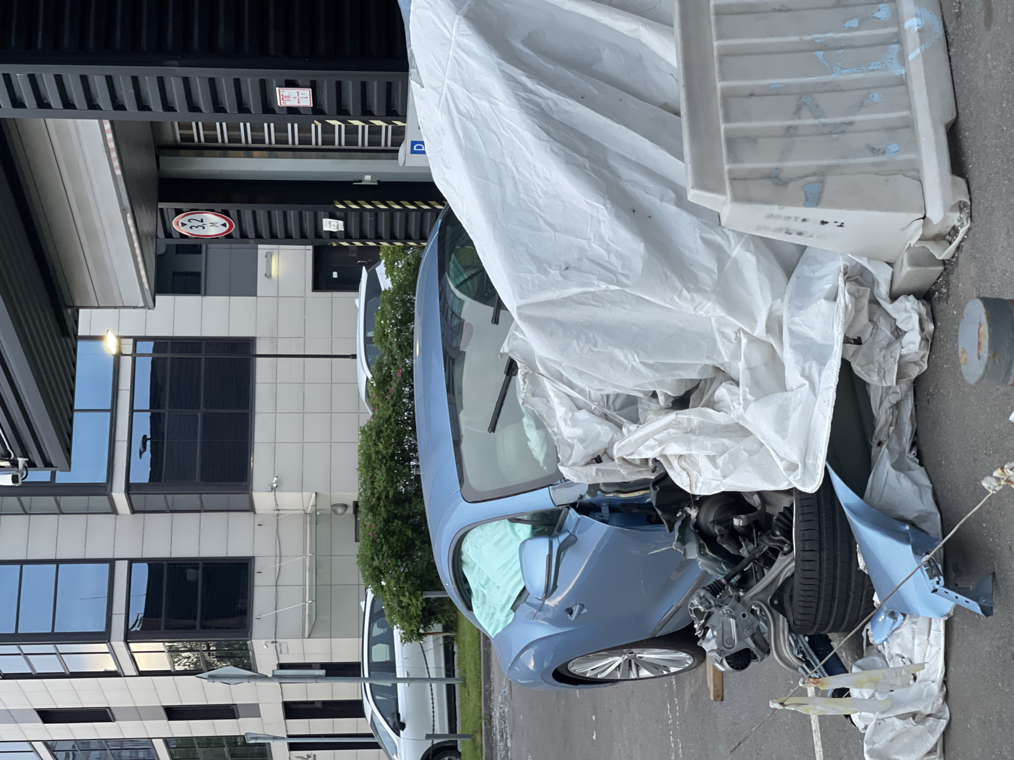 A blue car, heavily damaged with deployed airbags and tarpaulin covering the front, sits on an asphalt surface in front of a modern building. The building has a light-colored facade with large, reflective windows and an industrial-style loading bay with a height restriction sign indicating 3.2 meters. A white plastic barrier is positioned near the car. In the background, other vehicles and greenery are visible. The scene appears to be a street or parking lot in an urban environment.