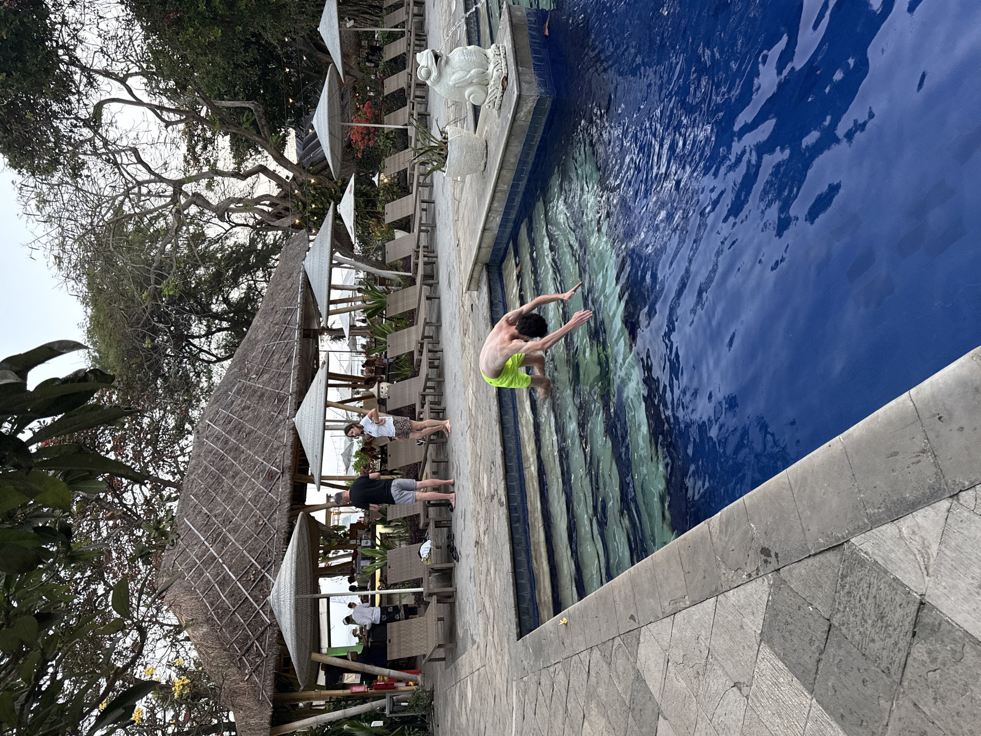 In Denpasar, Indonesia, a young person in neon green swim trunks dives into a blue swimming pool. The pool has tiered steps and is bordered by a stone patio. Behind the pool, lounge chairs and umbrellas are set up on a sandy area. Further back, a thatched-roof bar with patrons and staff is visible, and beyond that, lush tropical trees frame the sky. A decorative frog statue is placed near the pool's edge.
