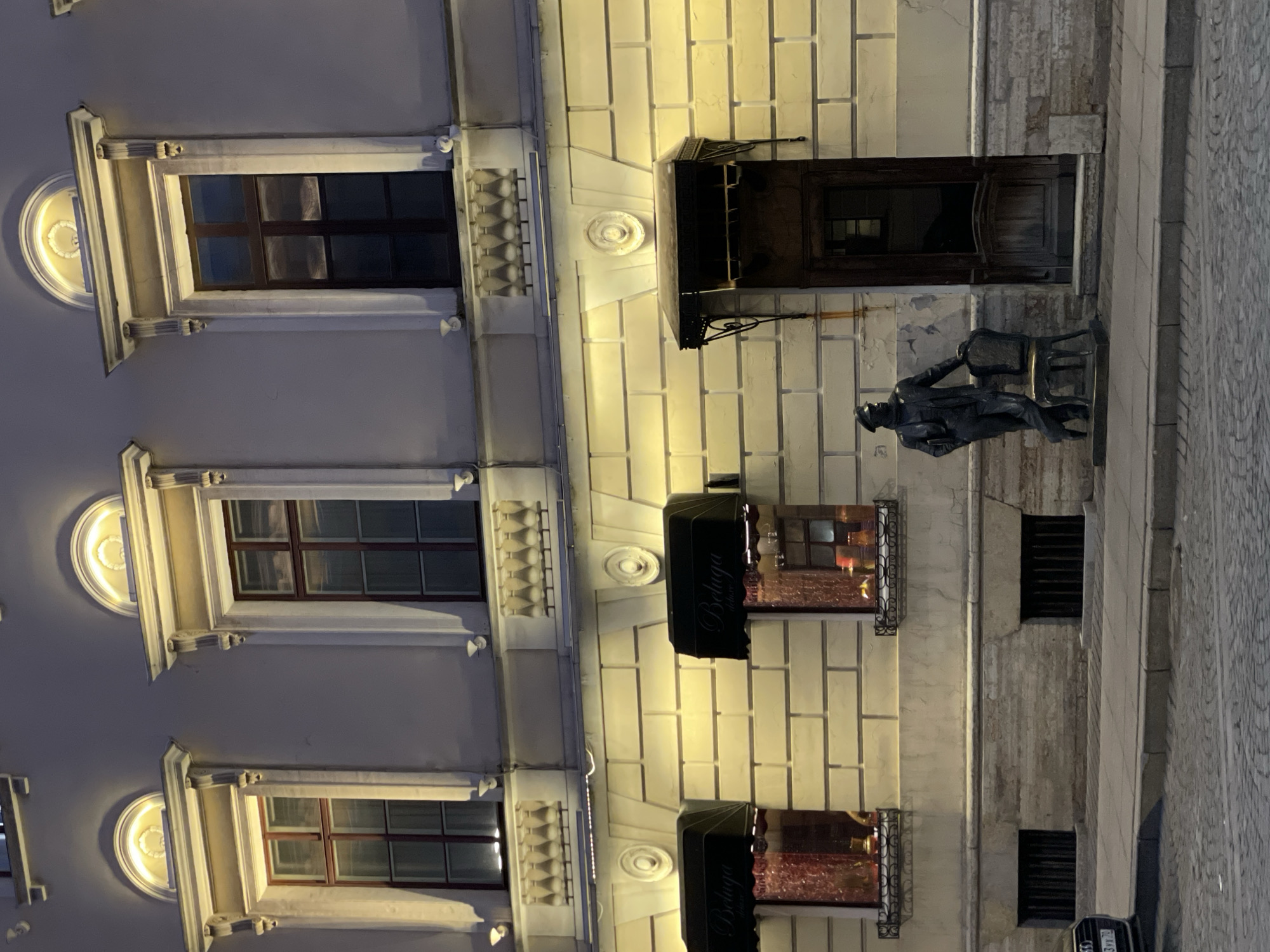 This media file captures a detailed night-time street scene in Saint Petersburg, Russia, focusing on the illuminated facade of a historic building and a bronze statue on the sidewalk. The building exhibits intricate architectural elements; the left section features a light-colored facade adorned with ornate window frames and semicircular decorative motifs above each window, housing glowing light sources. The windows themselves are dark-paned with visible reflections.
The right section of the building presents a different texture, composed of light-colored block-like stones or bricks, also warmly lit. Below a dark wooden door with a small canopy, a dark-colored statue of a man is prominently positioned on the cobblestone sidewalk. The man is depicted seated on a chair, wearing a hat and suit, with one arm resting on the chair's back. The chair features a distinctive golden or brass-colored frame.
Further along the building, two black awnings extend from the facade. Each awning clearly displays the text "Beluga deluxe" in white, suggesting the presence of an establishment such as a shop or restaurant. Below these awnings are decorative window boxes or small balconies with intricate metal railings, holding reddish-brown decorative elements. At street level, further to the right, are barred windows. A portion of a vehicle is visible at the far right, revealing a Russian license plate with the characters "943 УТ 78". The artificial, warm lighting suggests an evening or nighttime setting, highlighting the architectural details and creating a serene atmosphere.