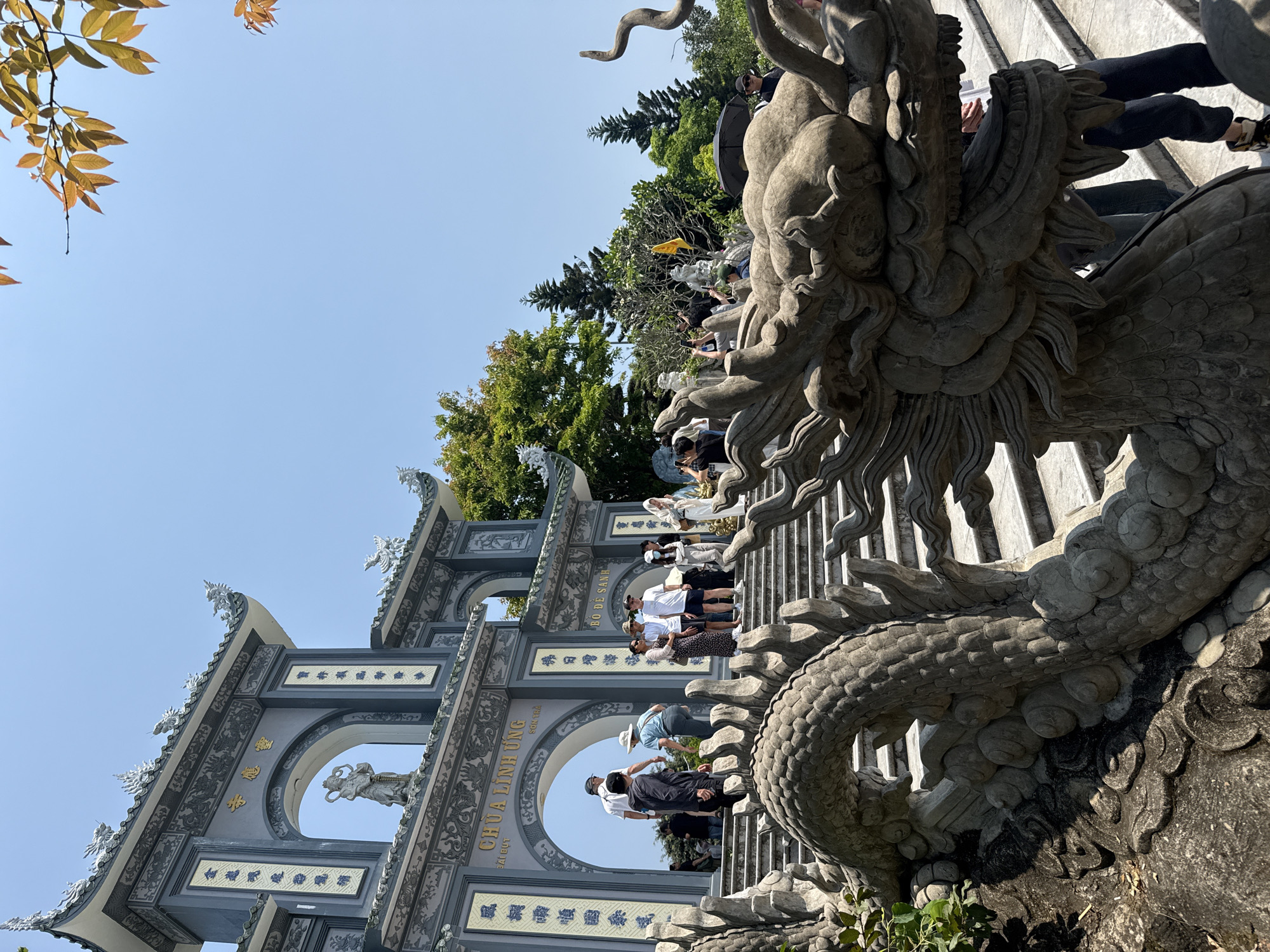 The image depicts a sunny outdoor scene at Linh Ung Pagoda on Son Tra Peninsula in Đà Nẵng, Vietnam, viewed from the base of a wide stone staircase leading to a large traditional gate. In the foreground, a prominent, elaborately carved stone balustrade shaped like a dragon forms the railing of the staircase, with its scaled body curving upwards and its head visible on the lower right. Numerous individuals, appearing to be tourists and visitors, are ascending the stone steps towards the gate. They are dressed in casual clothing, including t-shirts, shorts, dresses, and hats, with some holding phones or umbrellas. Some individuals are clustered on the steps, while others are in motion.
In the midground, the multi-tiered staircase terminates at a large, grey and white architectural gate with three arched entrances. Gold Vietnamese characters on the gate clearly identify it as "CHÙA LINH ỨNG SƠN TRÀ." The gate features intricate carvings, including dragon motifs along its roofline and a statue within the central archway. Beyond the gate and to the right, a dense collection of green trees, including pine-like species, covers a hillside. The sky is clear and bright blue, with some orange-brown leaves visible from an unseen tree in the upper right corner of the frame. A small yellow flag on a pole is visible amidst the trees on the right.