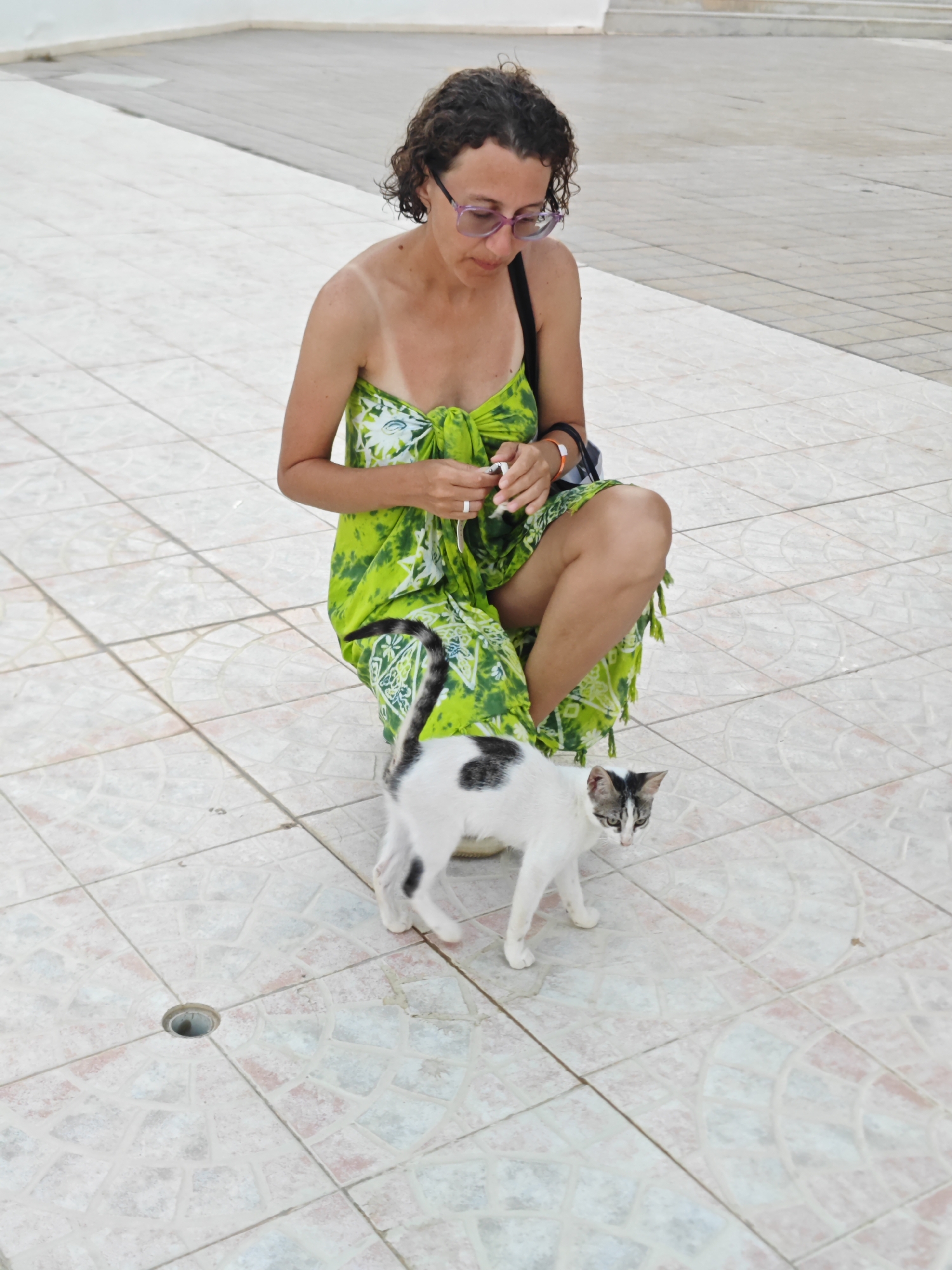 A woman with curly brown hair and purple-rimmed glasses is kneeling on a tiled outdoor surface in Hiboun, Tunisia. She is wearing a bright green and white patterned strapless dress that is tied at the bust and a dark strap from a bag is visible over her left shoulder. She appears to be holding something white and small in her right hand. A black and white cat with grey markings on its face is standing in front of her, looking at the camera. The ground is covered in pale pink and white square tiles arranged in a radiating pattern around a circular drain in the foreground. In the background, a lighter colored, flat surface slopes upward. The overall lighting suggests it is daytime.