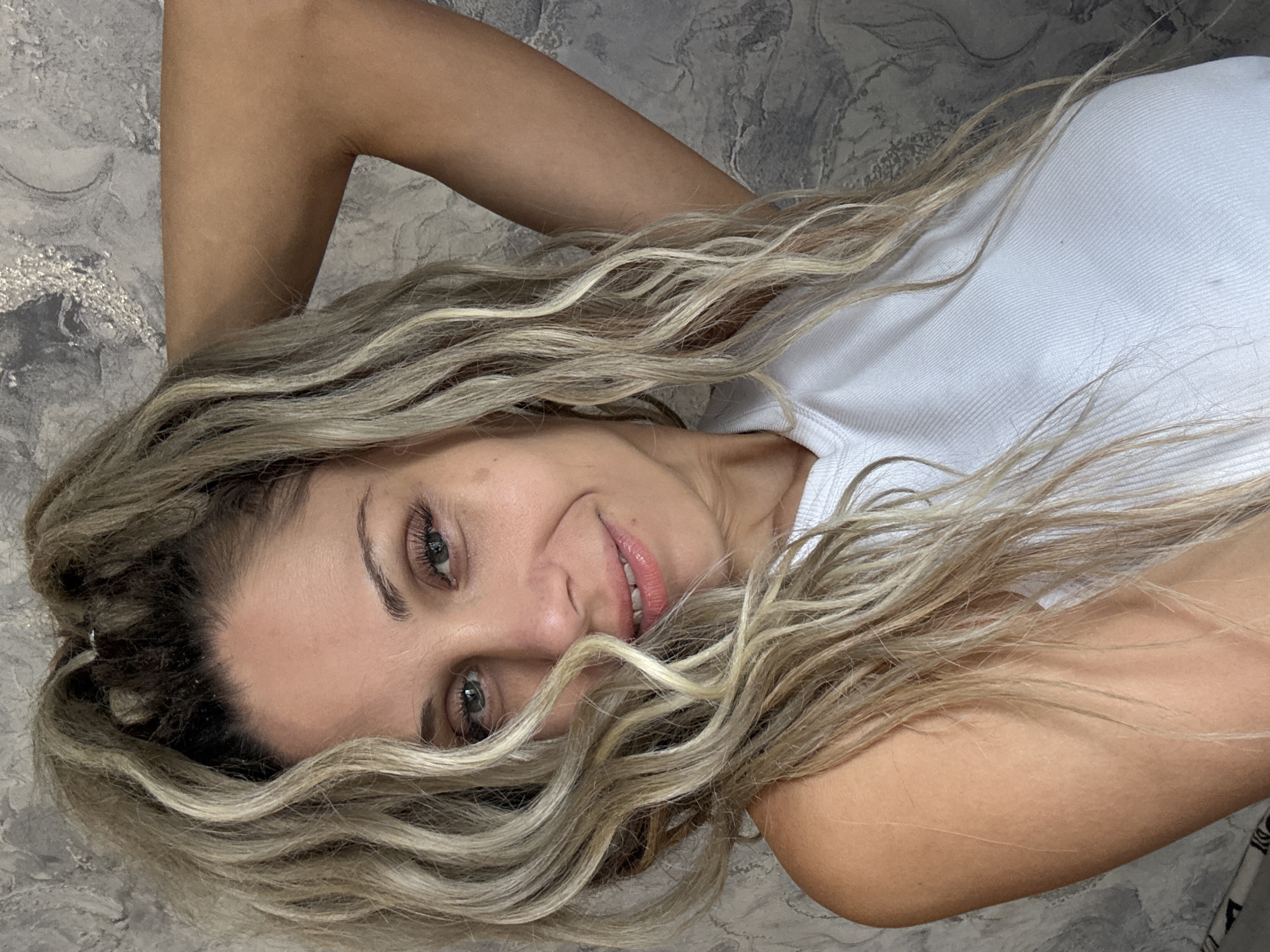 A woman with blonde, wavy hair is posing for a selfie against a gray, textured wall. She is wearing a white tank top and has her right arm raised behind her head, with her hand resting on her hair. Her eyes are green, and she has a slight smile on her lips. The lighting is natural, and the image is taken from the chest up. There is no indication of the location being Rusanovo, Russia, as no geographical markers are present in the image.
