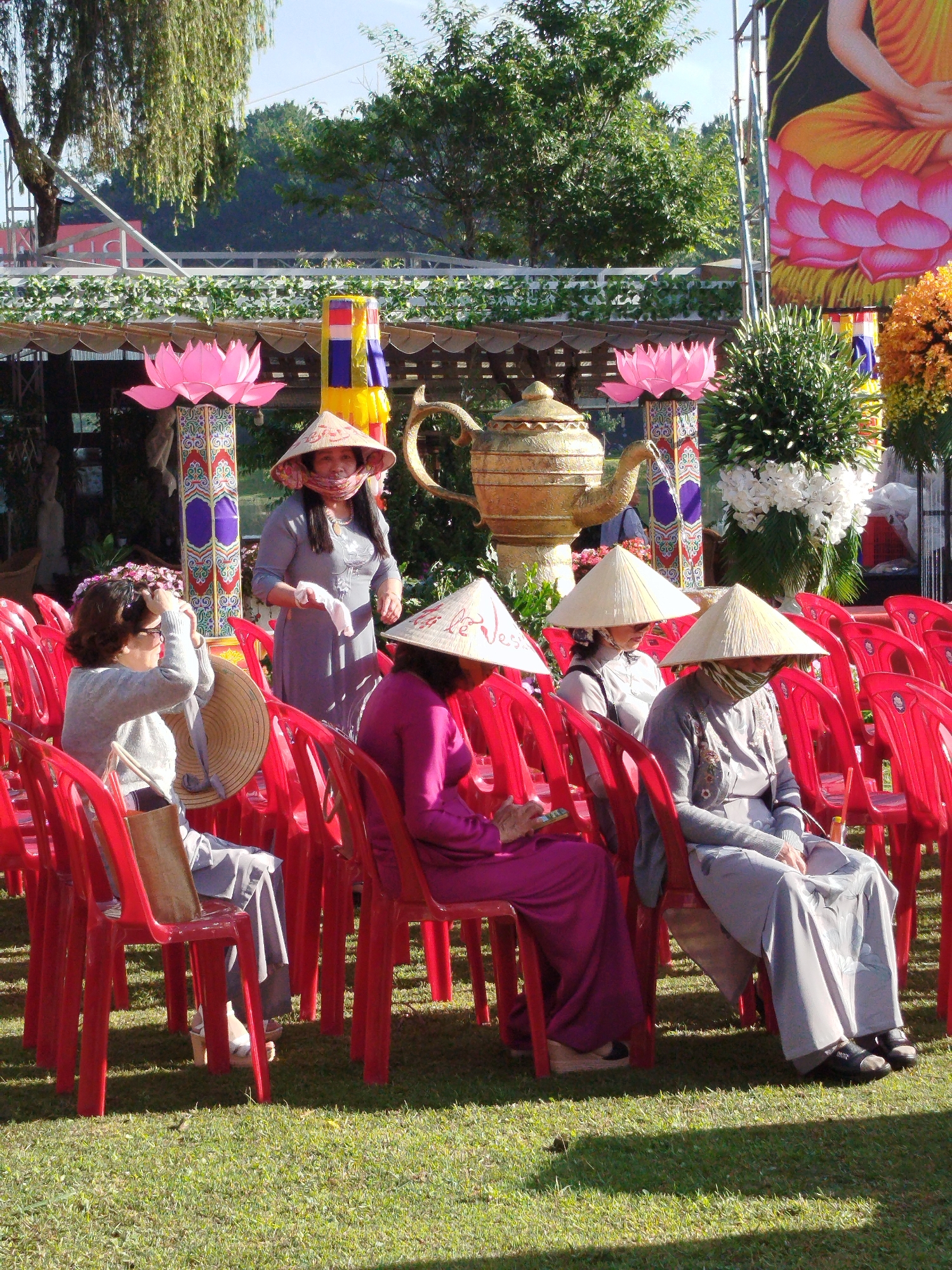 Several people are seated in red plastic chairs on a grassy lawn, wearing conical Asian hats and traditional garb, likely attending an event in Da Lat, Vietnam. One woman stands among the seated attendees, also wearing a conical hat and a face covering. Behind them, a decorative golden teapot and pink lotus flower sculptures are visible. A large banner depicting a seated Buddha is partially visible on the right side of the frame.