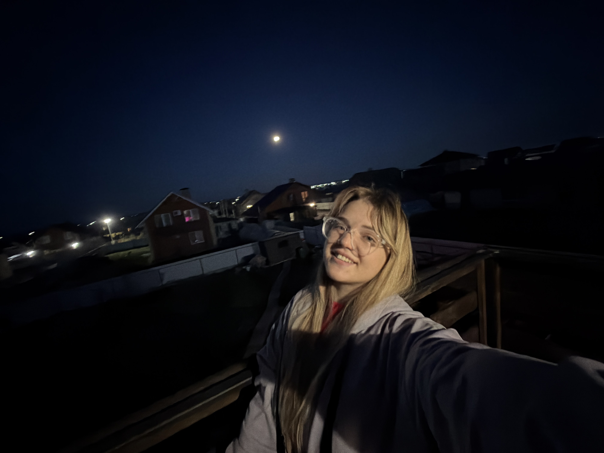 A woman with blond hair and glasses is smiling at the camera, taking a selfie. She is wearing a light gray jacket over a red shirt. In the background, there is a night sky with a full moon and houses. Street lights illuminate the scene. The woman appears to be standing on a wooden deck with railings. The photo was taken in the village of Большая Шильна, Russia.