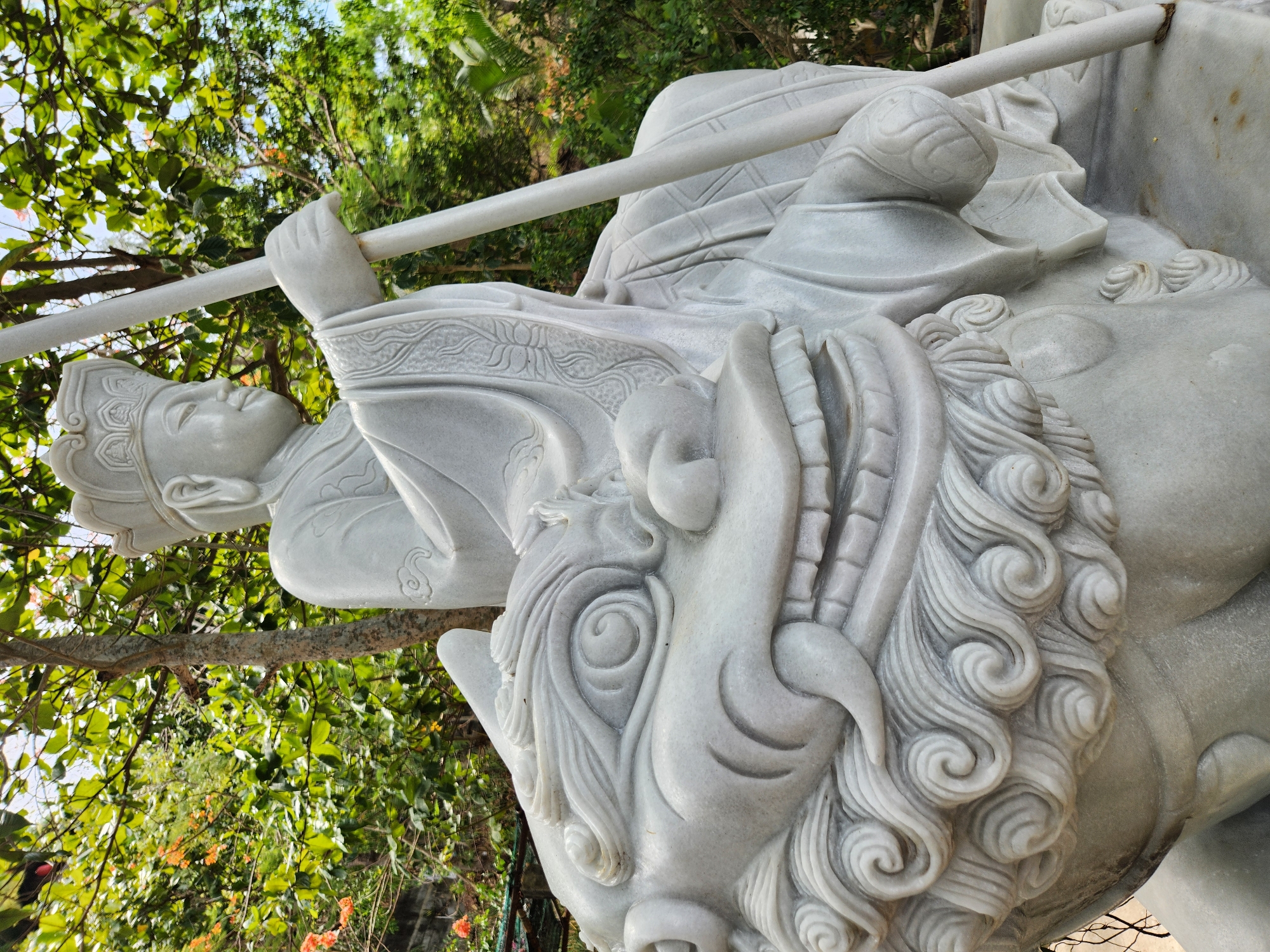 A close-up, eye-level shot captures a white marble statue of a robed figure wearing a headdress, seated atop a mythical beast. The figure's right hand grasps a long, thin staff. The beast's head, detailed with swirling mane, sharp teeth, and bulging eyes, dominates the foreground. The statue is situated outdoors, with lush green foliage and glimpses of orange flowers visible in the background. The lighting suggests it is daytime, likely under an overcast sky or in the shade of trees. This statue is located in front of the Non Nuoc Stone Carving Village, a renowned handicraft village in the Ngu Hanh Son district of Đà Nẵng, Vietnam, known for its marble sculptures.
