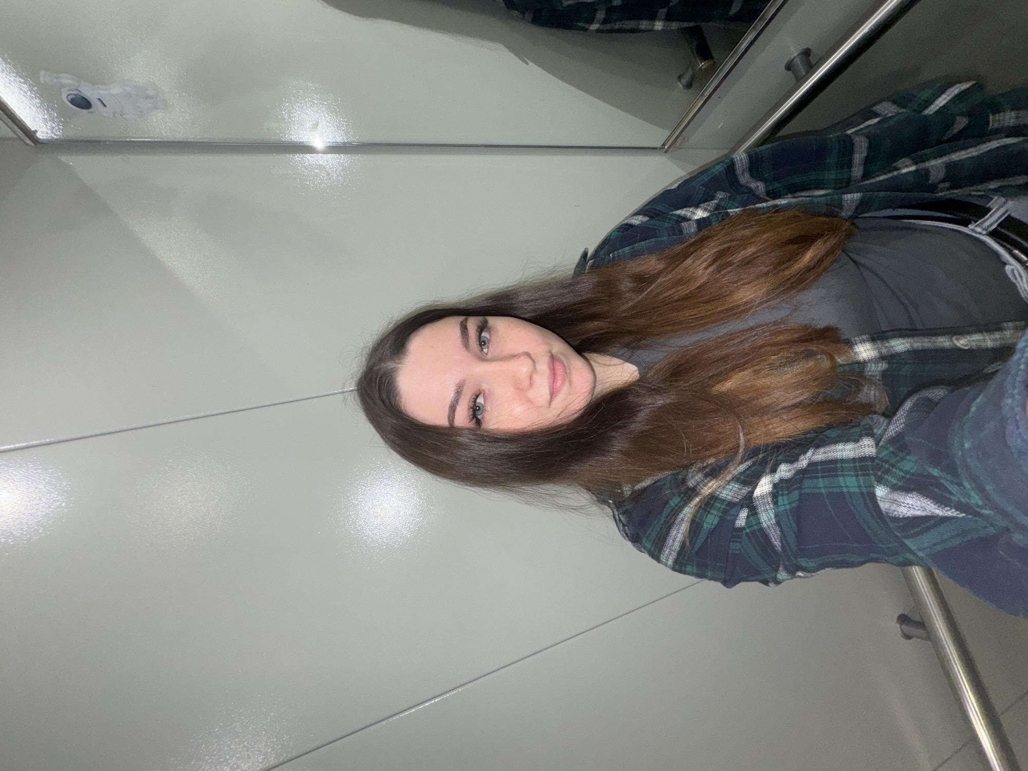 The image shows a young woman with long, brown hair, wearing a grey t-shirt and an open green and blue plaid flannel shirt. She has on false eyelashes and is looking directly at the camera with a neutral expression. She is standing in what appears to be an elevator, with grey metallic walls and a handrail. A mirror is visible on the right side of the frame, showing a reflection of the woman and an astronaut sticker on the wall. The overall lighting is dim and diffused. The specific location within the city of Perm, Russia cannot be determined from the visual information provided in the image.