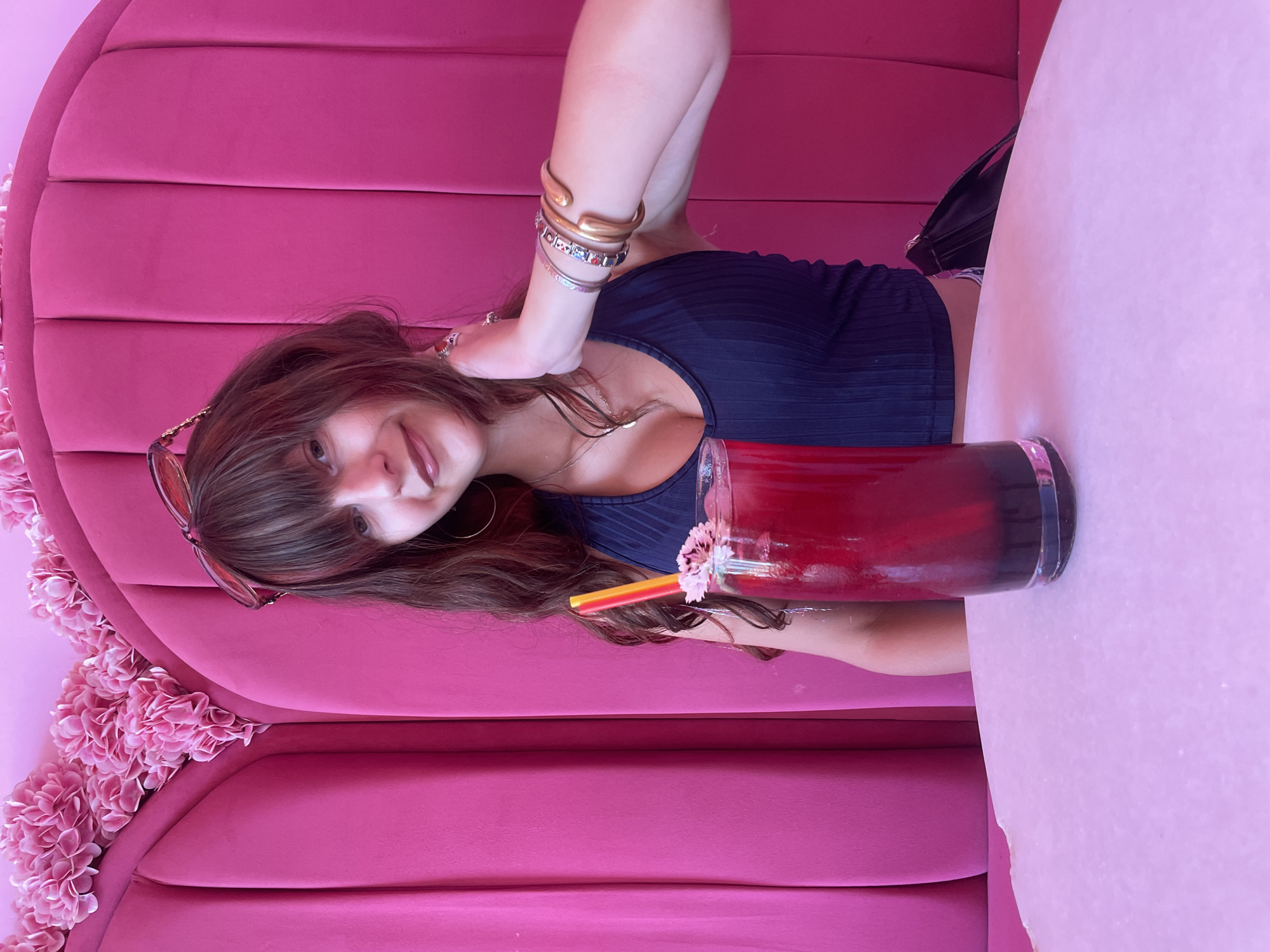 In Paphos Municipality, Cyprus, a young woman with brown hair and bangs is seated at a pink upholstered booth. She is wearing a dark blue ribbed tank top and has sunglasses perched on her head. She is adorned with several bracelets on her right wrist and a necklace. In front of her, on a pale pink table, is a tall glass filled with a dark red beverage. The drink has a yellow and orange straw and is garnished with a small pink flower. The background is dominated by bright pink upholstery and decorative pink flowers.