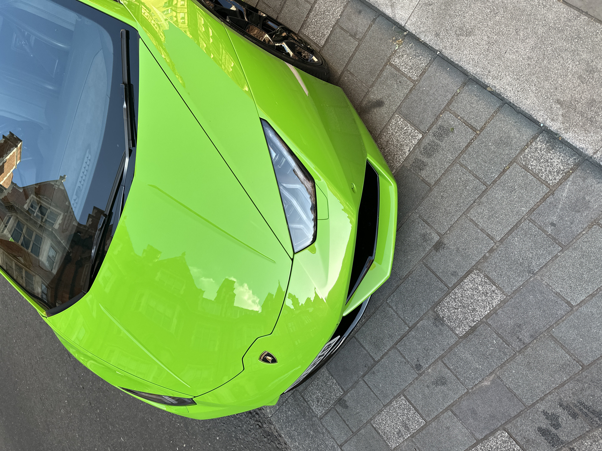 High-angle shot of a lime green Lamborghini parked on a cobblestone street in London, United Kingdom. The front portion of the car dominates the frame, with reflections of nearby buildings visible on its glossy surface. The license plate is partially visible. The street is made of gray and beige cobblestones.