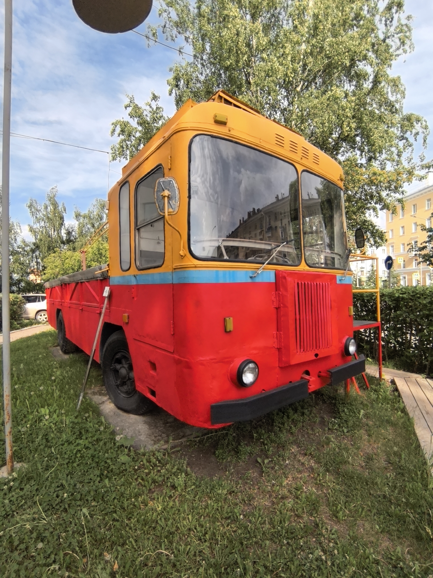 The image features a prominent, brightly painted vehicle, likely an old bus or specialized truck, as its central subject. The front half of the vehicle, including the cabin, is painted a vibrant yellow, while the lower body and chassis are a striking red. A thin blue stripe separates the yellow and red sections along the side. The vehicle has a large, multi-paneled windshield reflecting buildings and trees, large round headlights, and a sturdy black bumper. A vertical slatted grille is visible on the red front section below the windshield. A side mirror extends from the yellow cabin.
The rear section of the vehicle appears to be an open flatbed, partially covered by a dark tarp, and a white car is faintly visible behind it on the left. The vehicle is positioned on a raised, grassy mound, with lush green grass and some dandelions covering the ground around it. A tall, light-colored pole stands vertically on the left side of the frame. To the right of the vehicle, a yellow metal railing or ladder structure leads up to what looks like a small wooden platform or walkway. In the background, large trees with dense green foliage dominate the upper half of the image, and multi-story residential buildings, one predominantly yellow, are visible further back. A round, dark object hangs from above in the top center of the frame, partially obscured.
The scene is outdoors during the daytime, under a clear sky with scattered white clouds, suggesting good weather. There are no people visible, and no specific activity is taking place; the vehicle appears to be stationary, possibly on display or repurposed. No legible text is visible on the vehicle or in the surroundings.