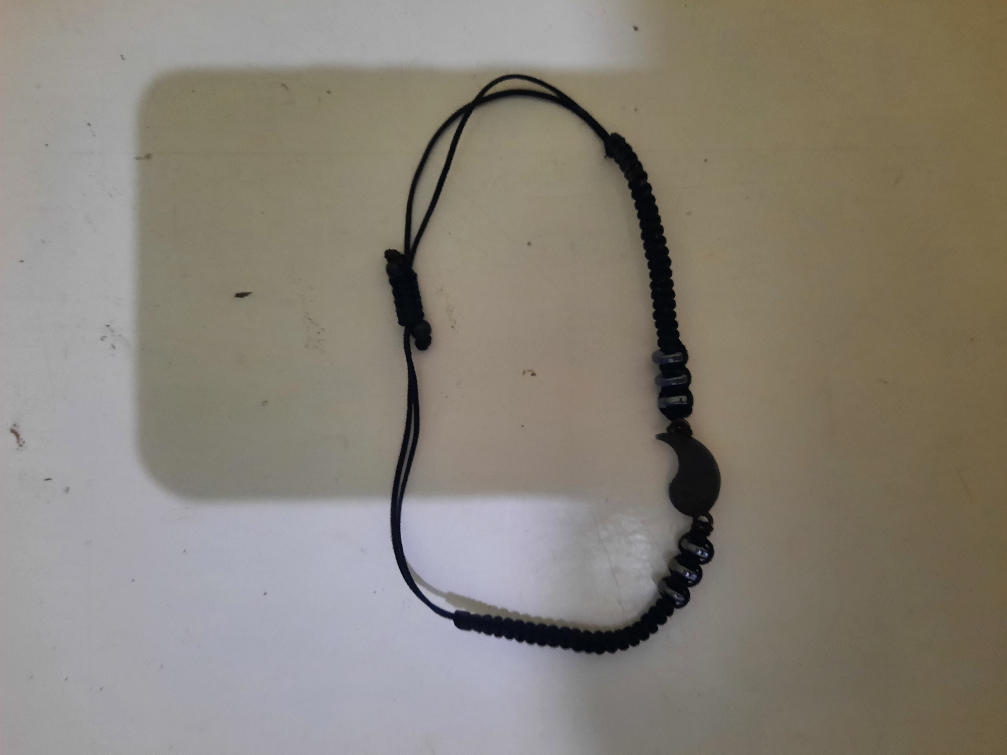 The image shows a close-up, overhead view of a black woven bracelet with a pendant. The bracelet is on a light-colored, textured surface, possibly a table or countertop. The bracelet itself is made of black cord, with a section of tightly woven cord on the left side, and adjustable sliding knots on the right. There are several small, dark, cylindrical beads spaced along the bracelet. Centered on the bracelet is a black, teardrop-shaped pendant with a curved line, resembling a paisley or stylized comma shape. The lighting in the image creates a soft shadow on the upper right portion of the surface. The image does not depict any elements that would suggest a specific location in Lhokseumawe, Indonesia.