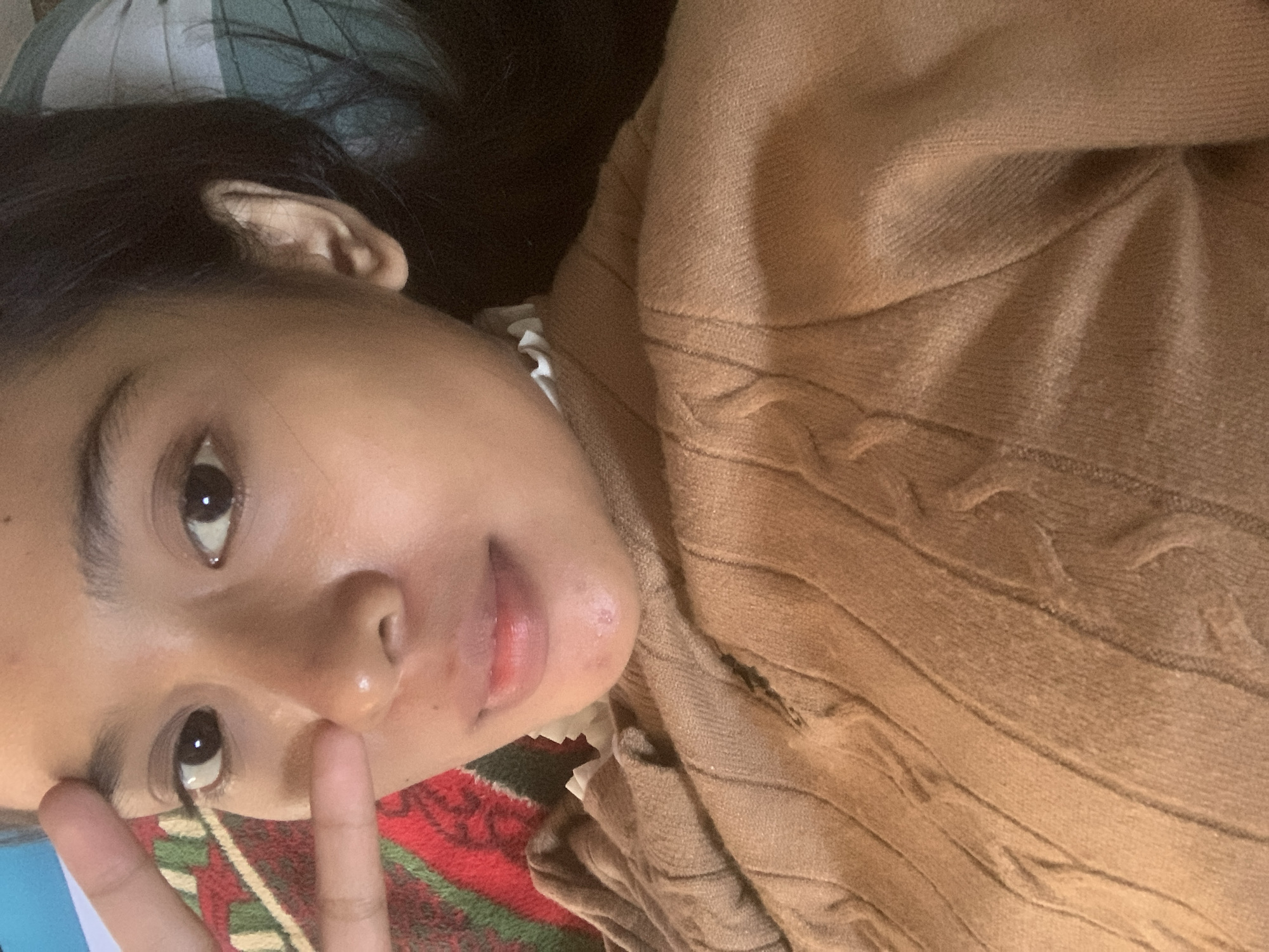 A close-up, overhead shot shows a person lying down and looking up at the camera. The person has dark hair, brown eyes, and appears to be wearing a tan sweater. Their right hand is raised, with two fingers extended in a peace sign gesture near their forehead. A red and green patterned blanket is visible behind their head and left shoulder. The background also includes a pillow with a green and white checkered pattern. The person's skin appears to have some minor blemishes on the chin.