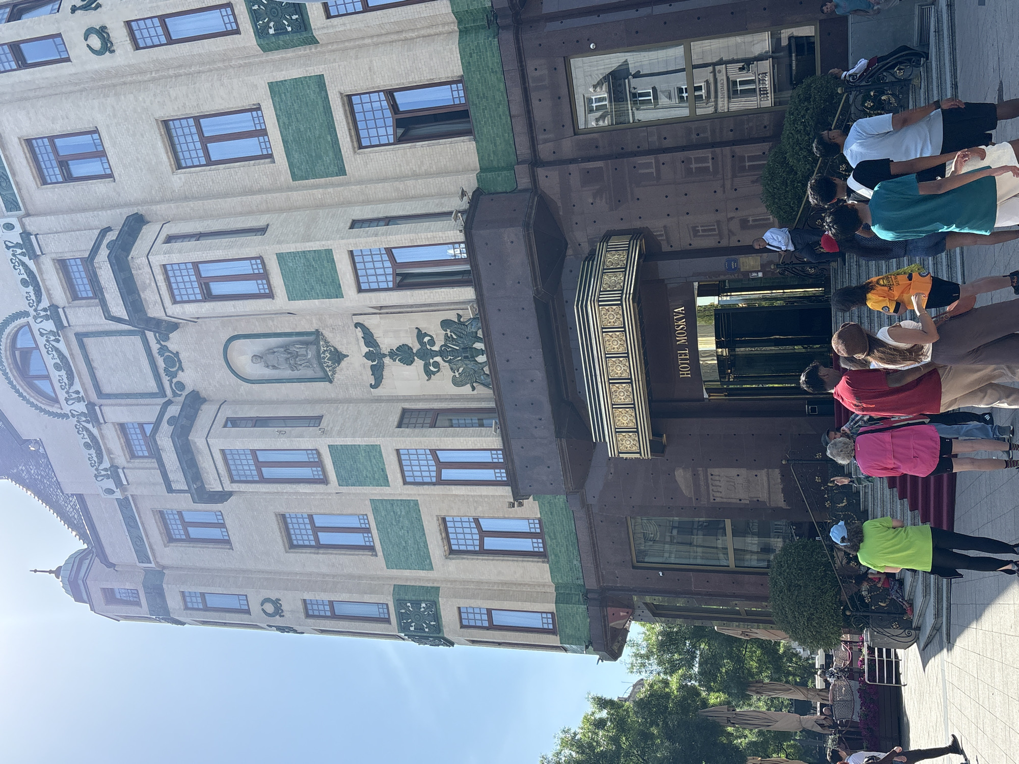The image depicts the facade of Hotel Moskva in Belgrade, Serbia, with a group of people standing near the entrance. The hotel has a distinctive architectural style with light-colored walls, green accents, and decorative details. "HOTEL MOSKVA" is visible above the entrance. The people appear to be a mix of ages and are dressed in casual clothing. Some are standing, others are walking. The weather seems sunny, and the sky is clear. Trees and outdoor seating are visible in the background, suggesting a public space or terrace area adjacent to the hotel.
