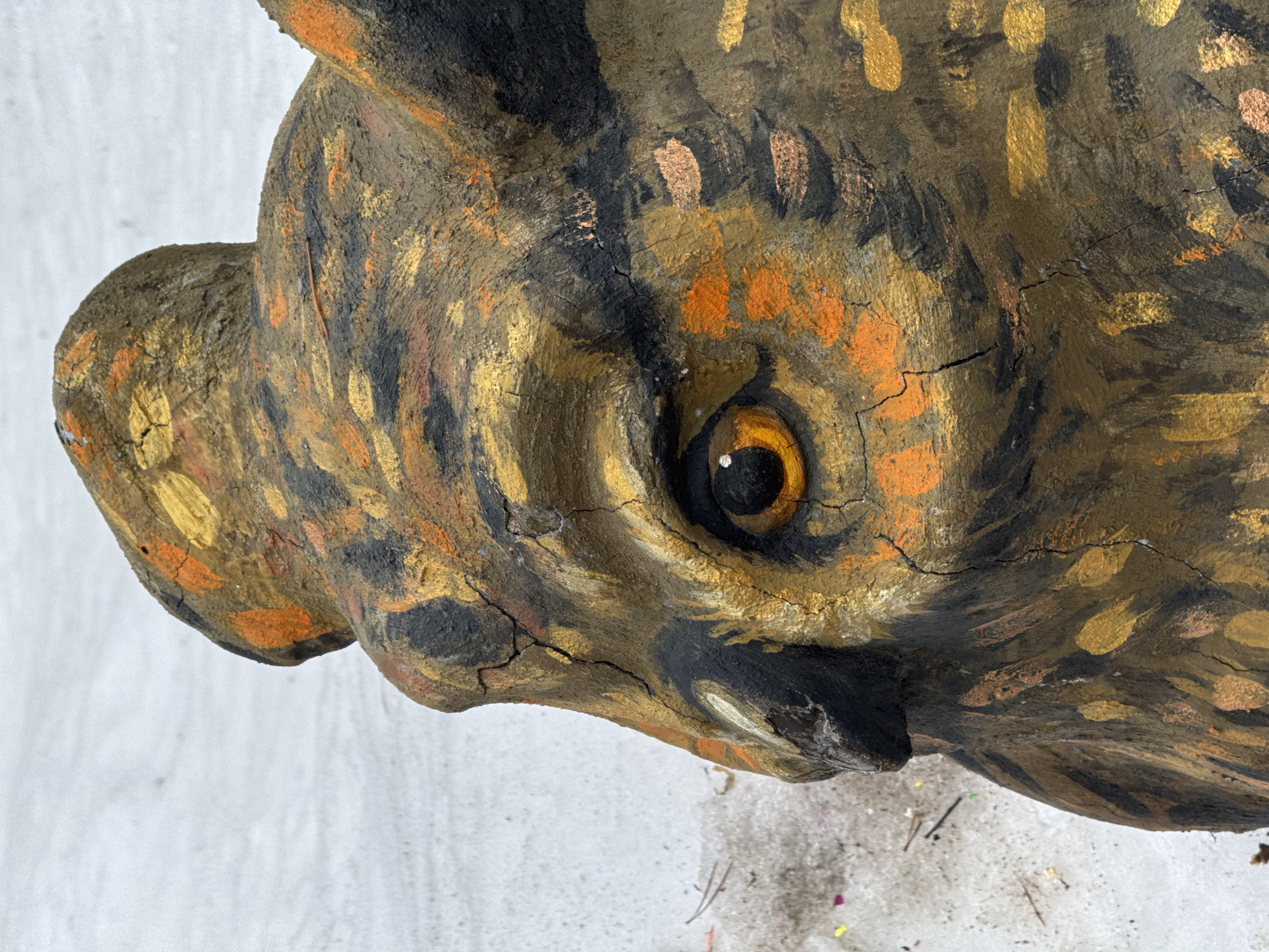A close-up, high-angle shot reveals a painted, possibly ceramic or papier-mâché, wild boar head. The head is primarily painted in earth tones: browns, blacks, and gold. The painting style is somewhat rough, with visible brushstrokes. Cracks are present in the paint and surface material, suggesting age or fragility. The boar's eye is a focal point, painted with a dark center and amber iris. The background is a blurred white surface. The image does not confirm its origin in Chaikovsky, Russia, as there are no discernible landmarks or identifying features. There are no people or actions visible in the image.