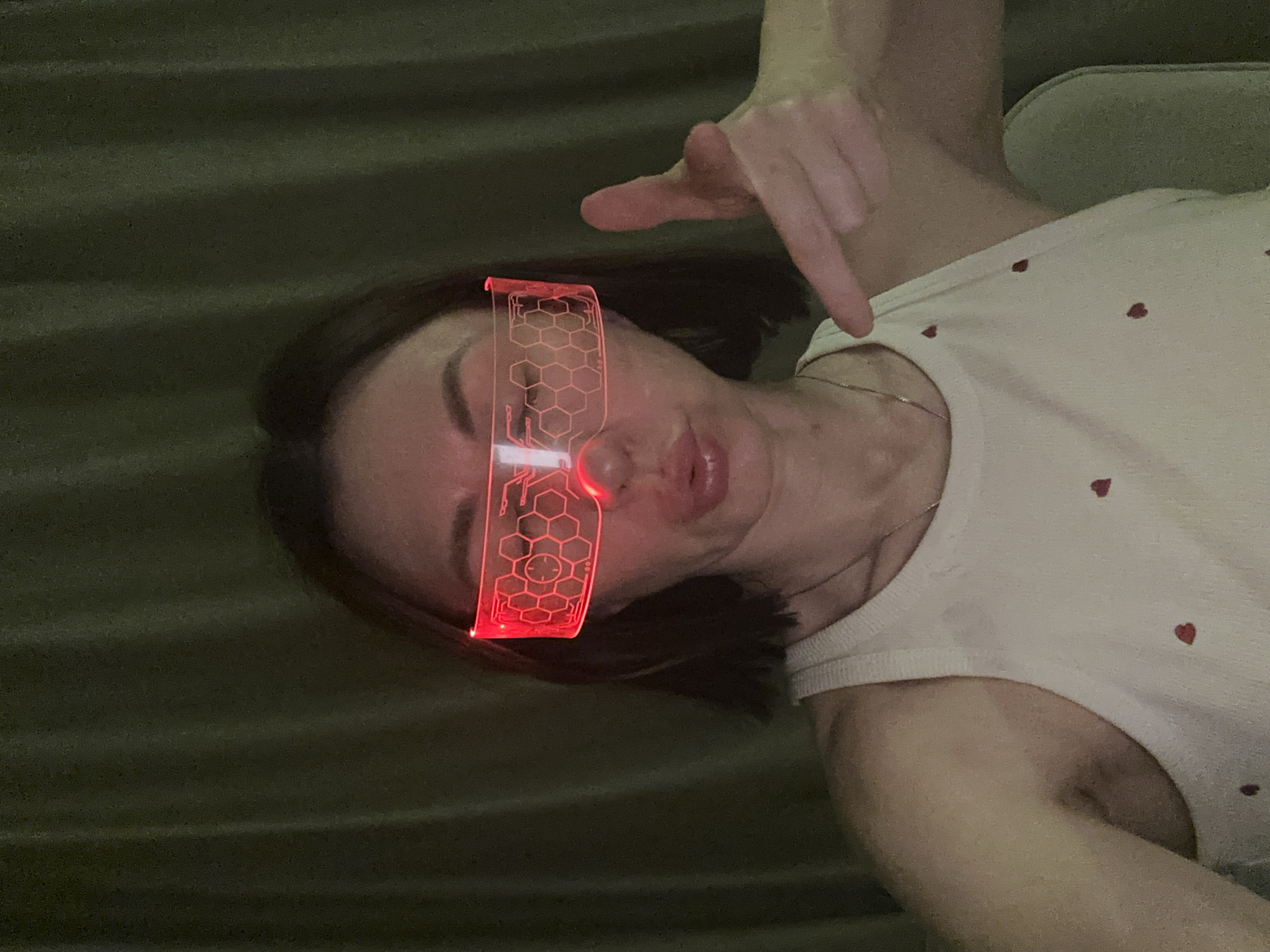 A person wearing red light-up glasses is lying on a green surface, possibly a bed or blanket. The person is wearing a white shirt with small red heart patterns and has their hand raised with their finger pointing down. A silver chain necklace is visible around their neck. There is no evidence of a location in Votkinsk, Russia.
