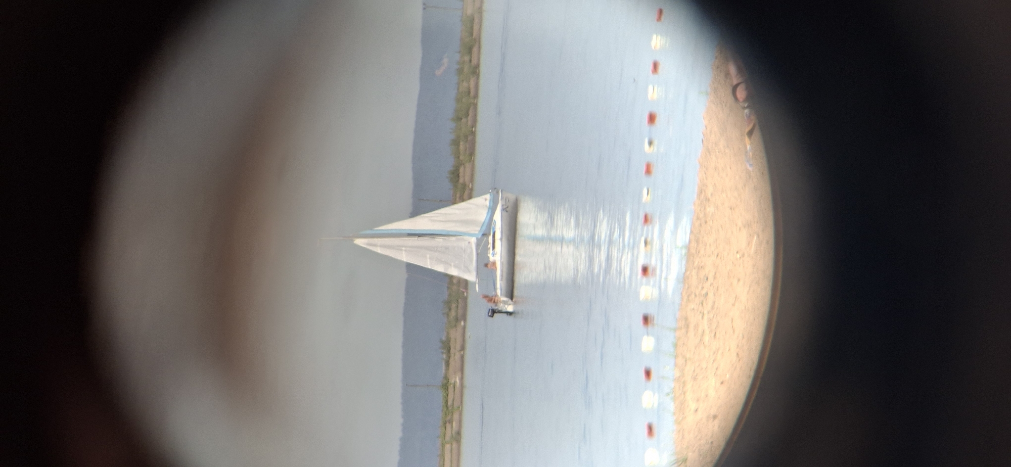 The image displays a daytime scene of a sailboat on a large body of water, likely the Votkinsk Reservoir, viewed through a circular optical instrument, such as binoculars or a telescope, from a sandy shoreline in Chaikovsky, Russia. The sailboat, featuring a white hull and white sails with distinct light blue trim, is positioned towards the center of the frame, moving left to right. An outboard motor is mounted at its stern. Two individuals are visible on the boat: one person is seated on the port side near the stern, and another is seated further forward in the cockpit.
In the immediate foreground, a sandy beach transitions into the water, where a line of red and white buoys marks a boundary. The water surface is calm, showing reflections. The background consists of distant land with a visible treeline or dense vegetation, and low hills under a hazy, light gray sky. The overall impression is a leisurely outdoor scene on a moderately clear day.