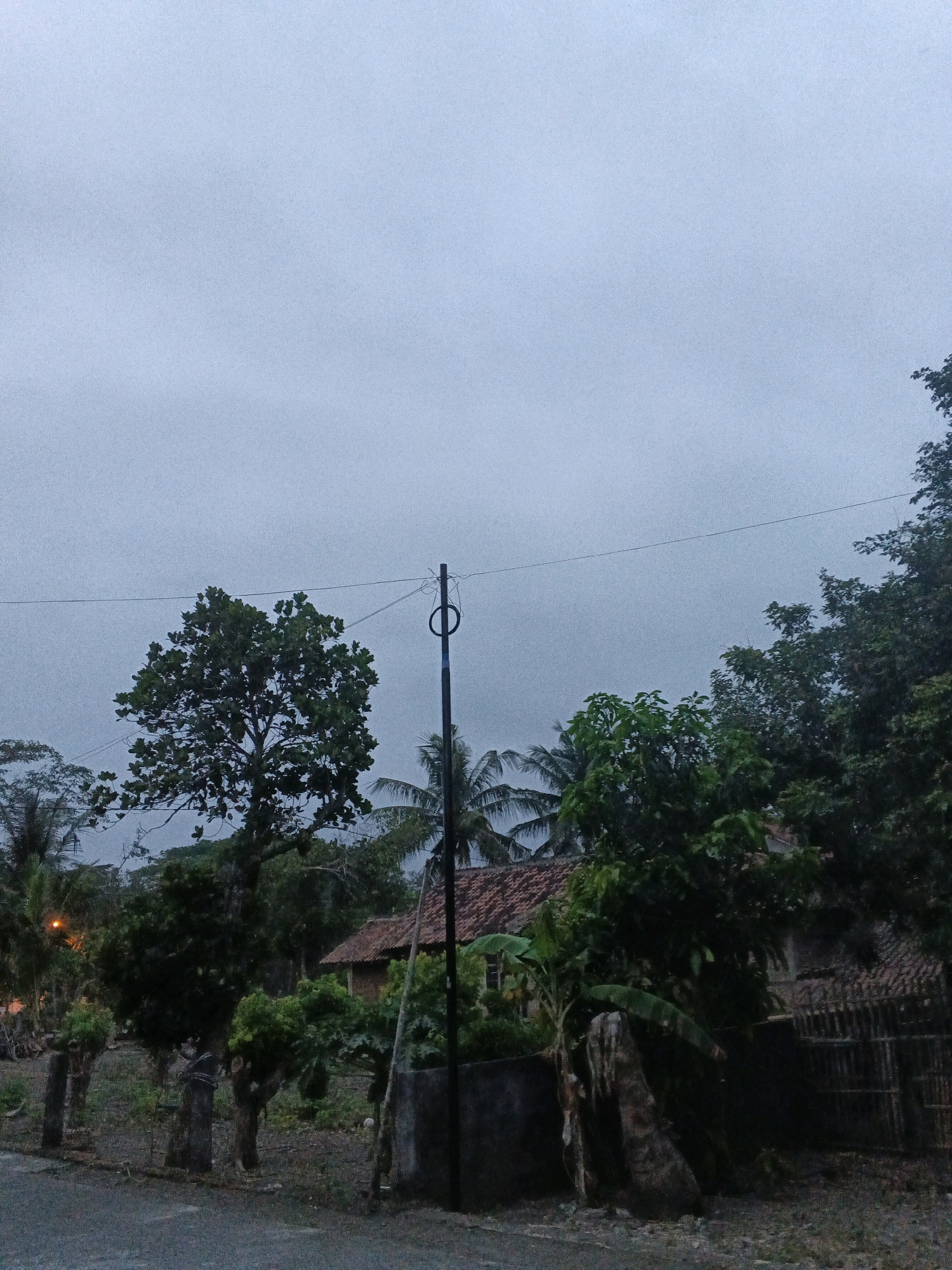 This image captures a cloudy, overcast sky over a rural scene in Wates, Indonesia. A dark, thin utility pole stands in the center, with a wire stretching horizontally across the frame. To the left of the pole, a large tree with dense green foliage dominates the middle ground. Behind it, the terracotta-tiled roof of a building is visible, peeking through the trees. To the right of the pole, more trees and vegetation are present, with a traditional bamboo fence visible in the far right. The foreground shows a dirt or gravel path and some uneven terrain with scattered debris. The overall atmosphere is dim and muted, suggesting a pre-dawn or post-dusk setting, or possibly a heavily overcast day.